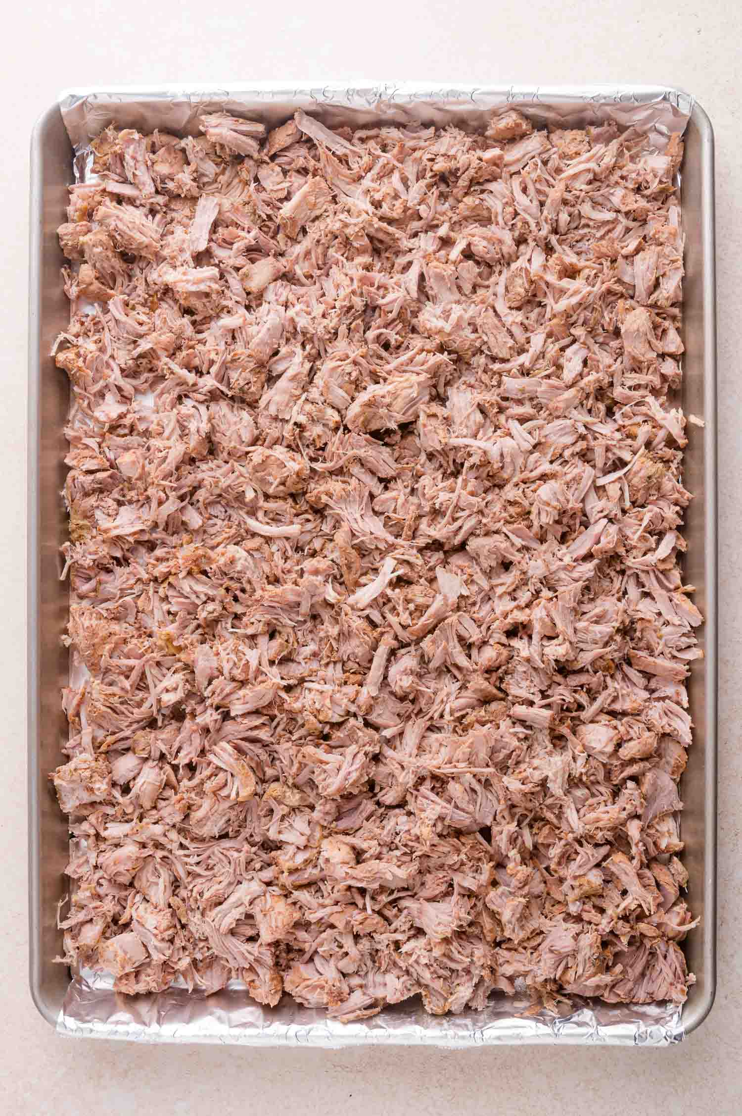 A baking sheet lined with foil is filled with a large amount of shredded cooked pork spread evenly across the surface.