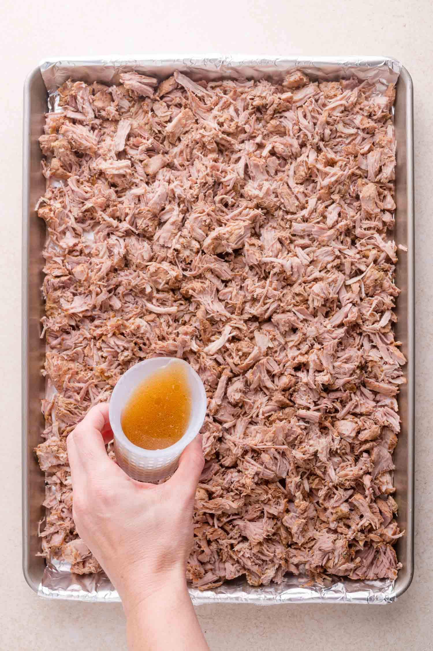 A hand pours broth from a cup over a baking sheet covered with shredded cooked pork.