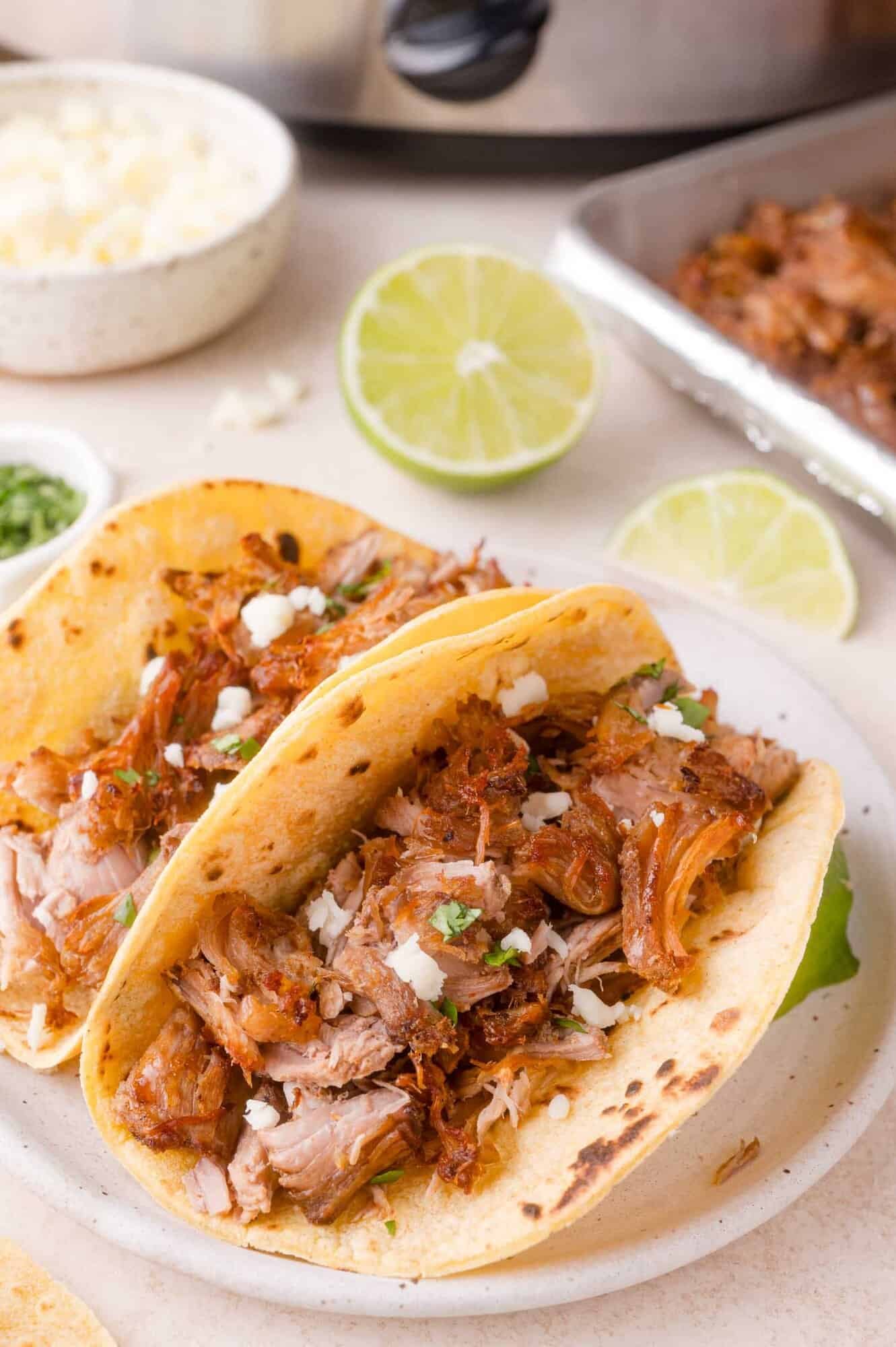 Two carnitas tacos with shredded pork, crumbled cheese, and chopped herbs on corn tortillas, served on a plate with lime halves and a tray of meat in the background.