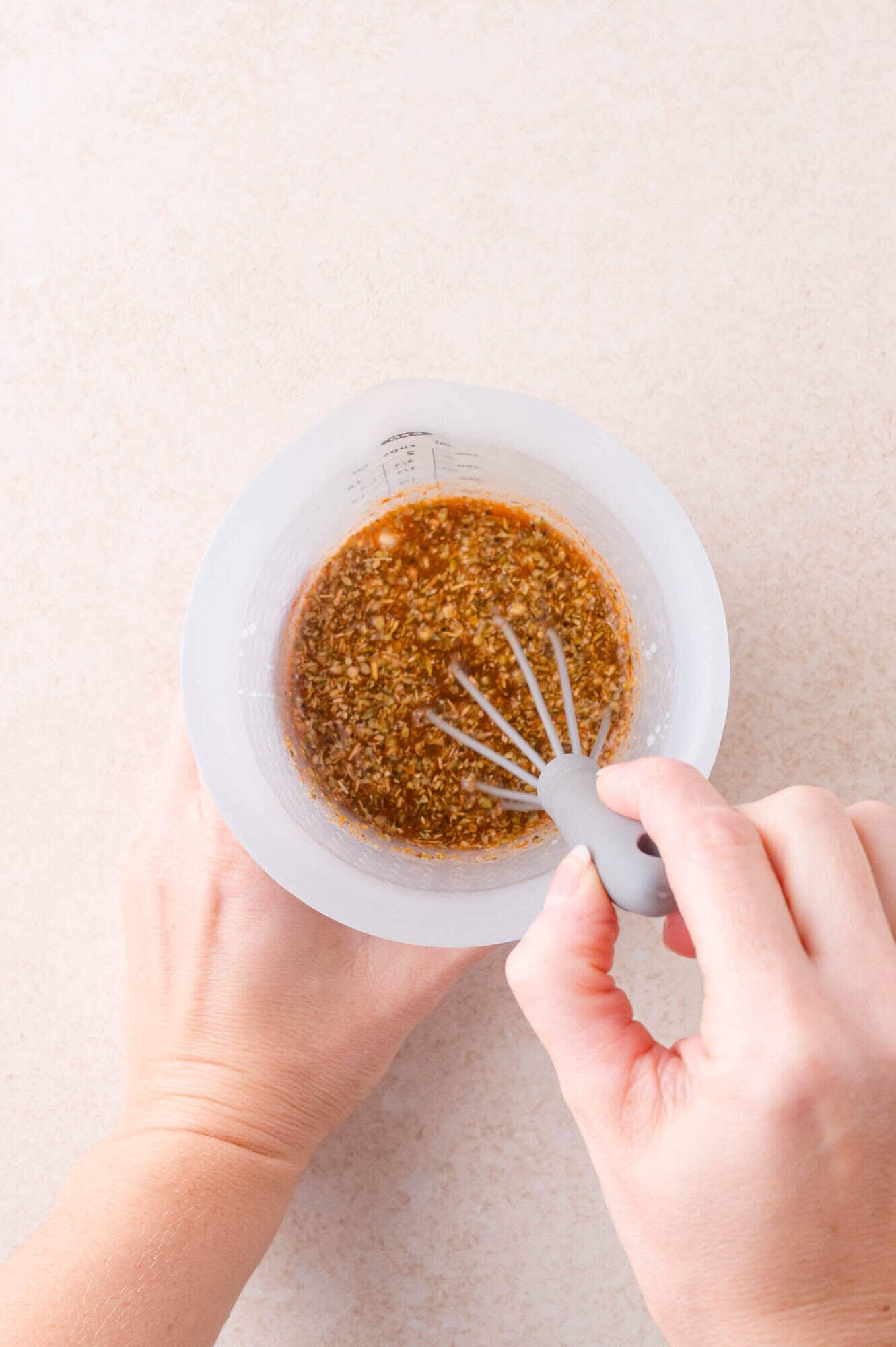 A person uses a small whisk to mix a marinade or sauce with visible minced ingredients in a clear measuring cup on a light surface.