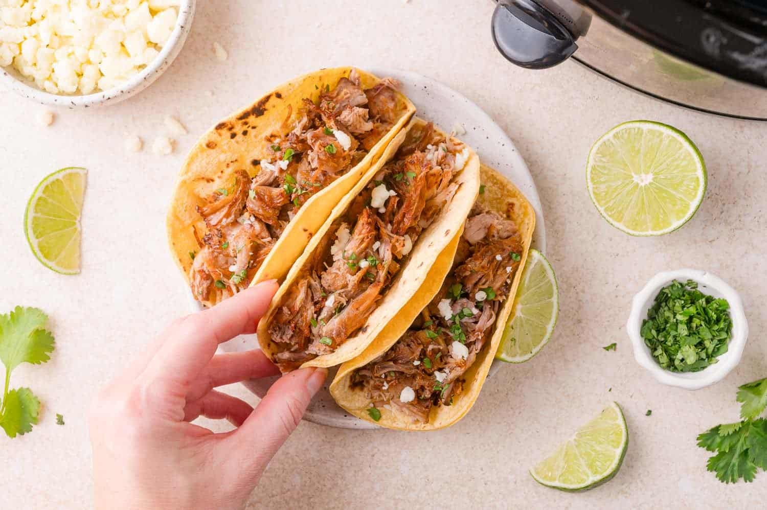 Hand holding a taco with shredded carnitas, cheese, and herbs; another taco on the plate; surrounded by lime wedges, cilantro, cheese, and a slow cooker.