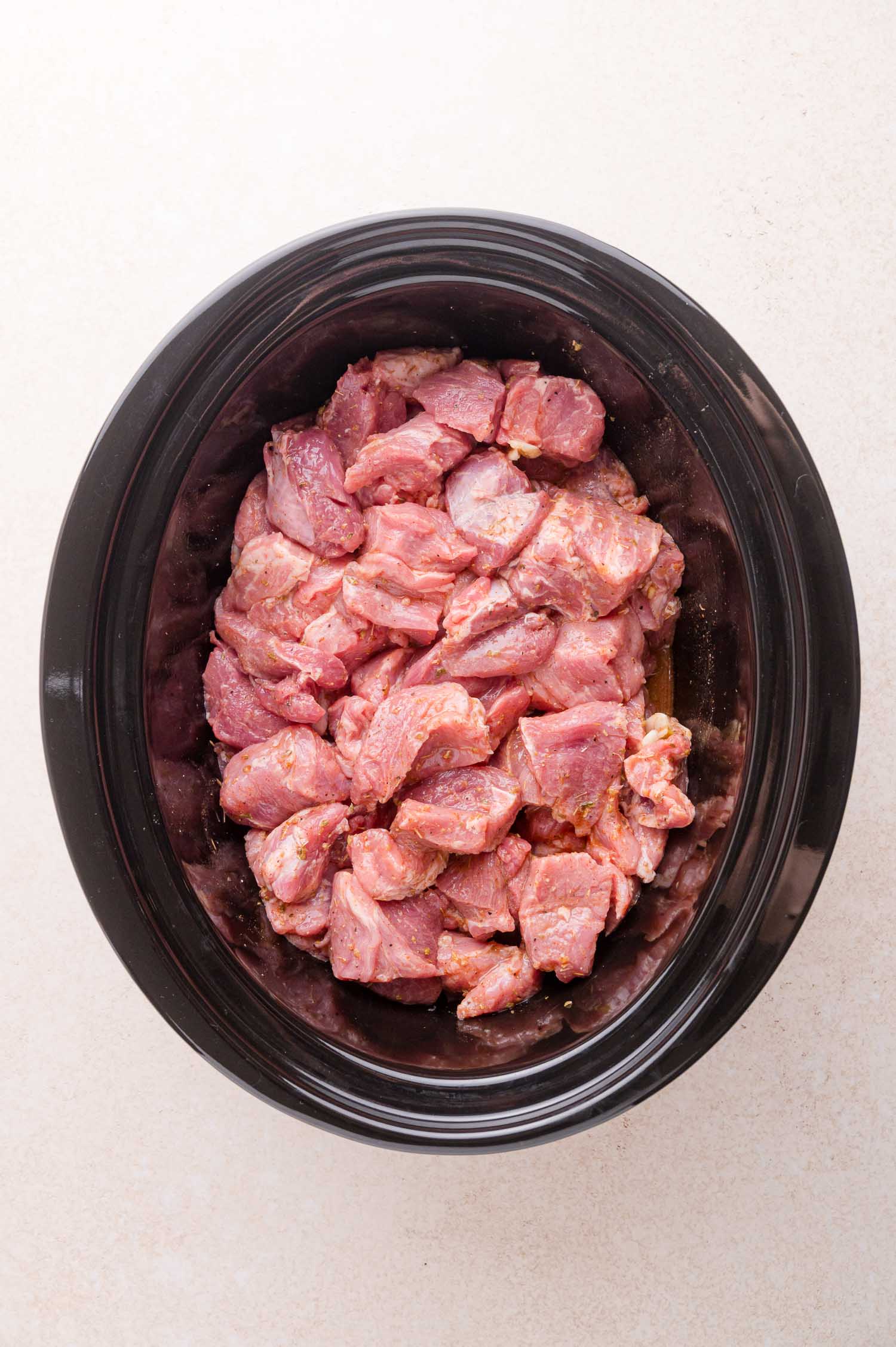 Chunks of raw, seasoned pork are placed inside a black oval slow cooker on a light-colored surface.