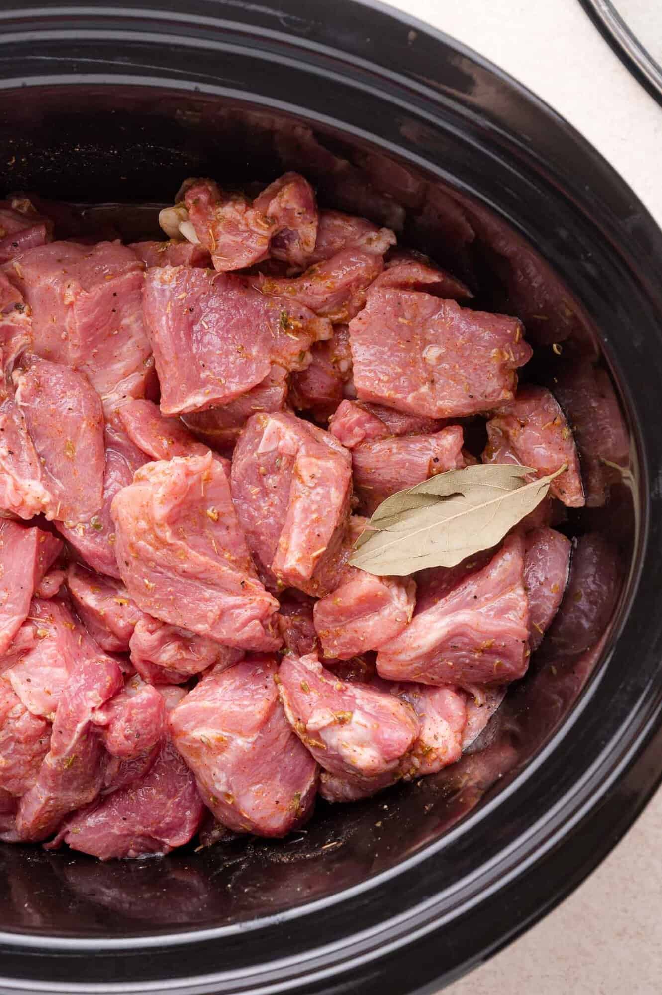 Chunks of raw, seasoned pork and a bay leaf in a black slow cooker, ready to be cooked.