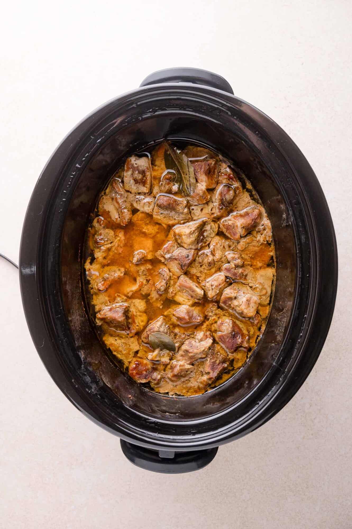 A slow cooker containing chunks of meat simmering in a brownish sauce with visible bay leaves.