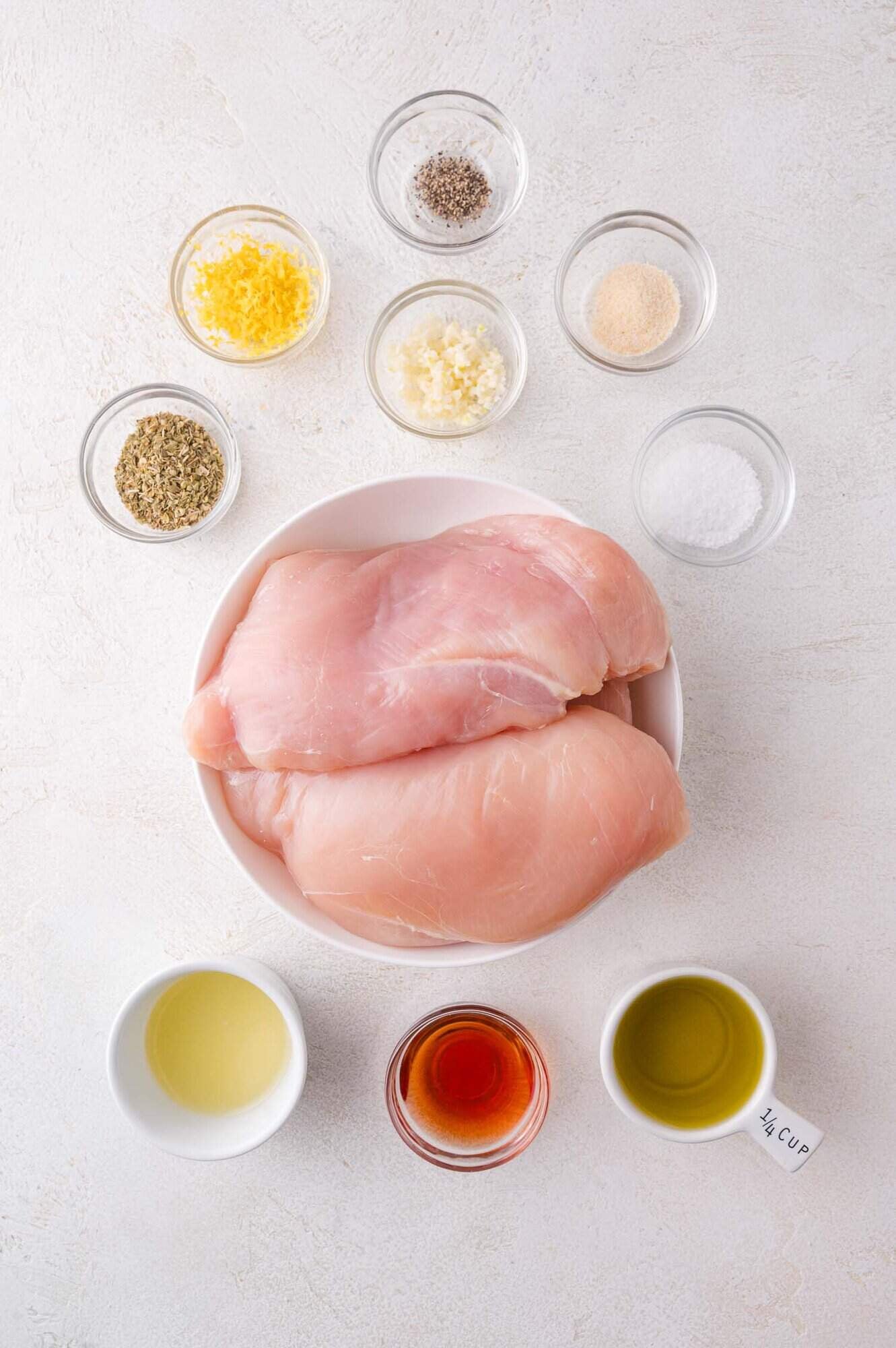 A bowl with two raw chicken breasts is surrounded by small bowls containing spices, oil, vinegar, minced garlic, salt, pepper, and lemon zest on a light surface.