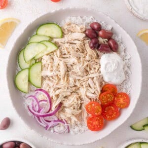 A bowl of shredded chicken on rice, topped with sliced cucumber, cherry tomatoes, red onion, olives, and a dollop of white sauce; surrounded by small bowls of ingredients.