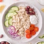 A bowl of white rice topped with shredded chicken, sliced cucumber, cherry tomatoes, kalamata olives, red onion, and a dollop of tzatziki sauce.