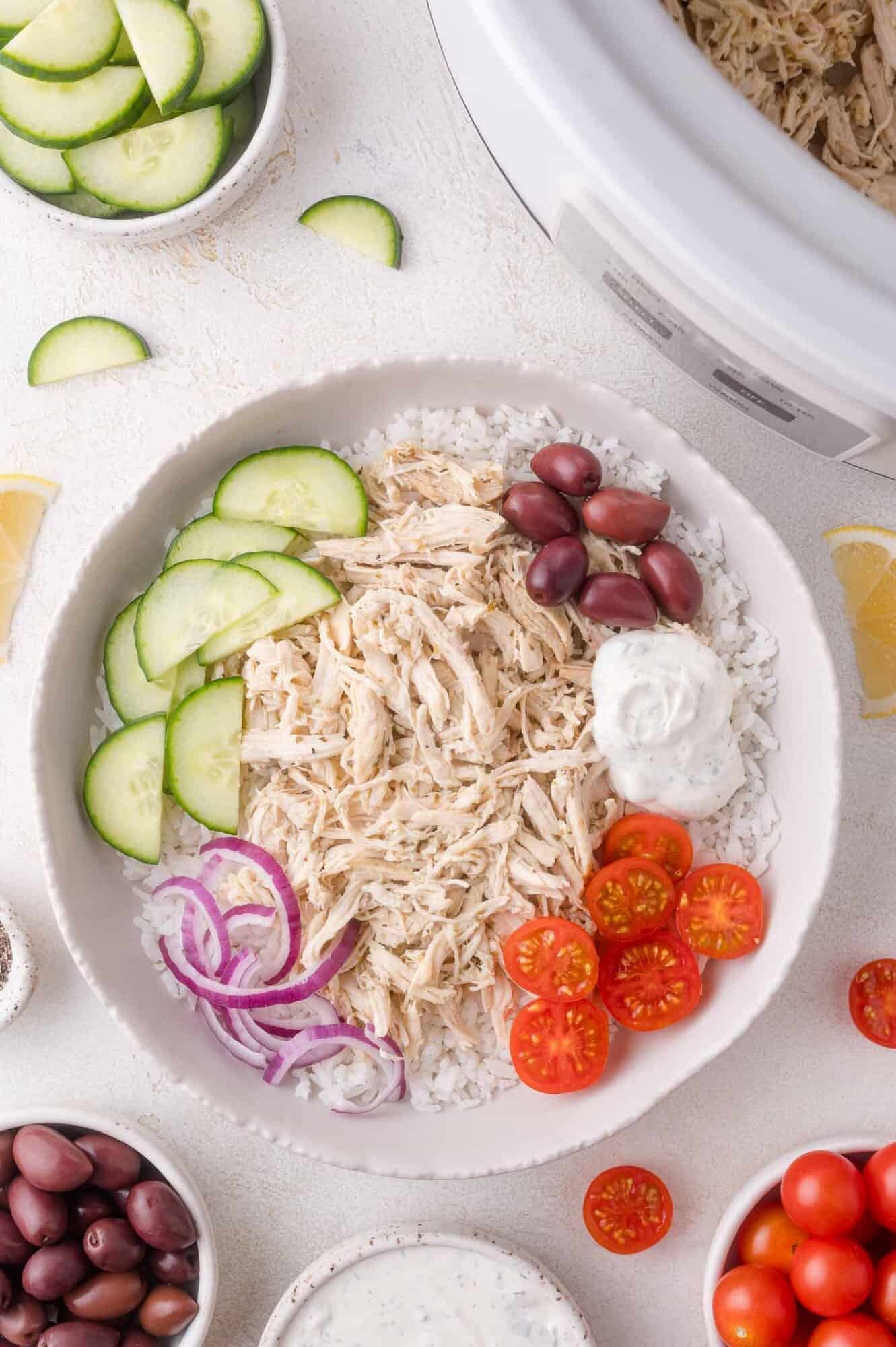 A bowl of shredded Greek chicken, sliced cucumbers, cherry tomatoes, red onions, kalamata olives, and tzatziki sauce on rice, surrounded by bowls of extra ingredients and a slow cooker.