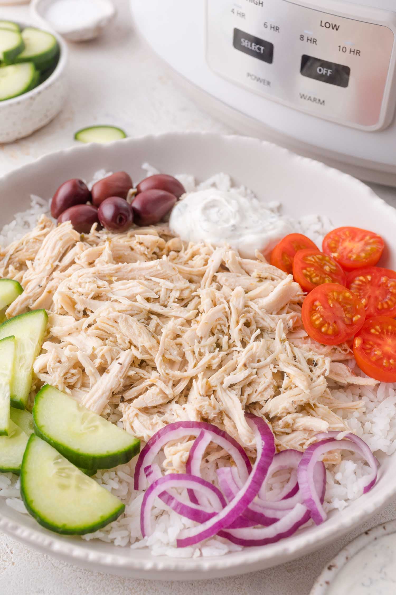 A bowl of shredded chicken on rice, topped with sliced cucumbers, cherry tomatoes, red onions, kalamata olives, and a dollop of white sauce.