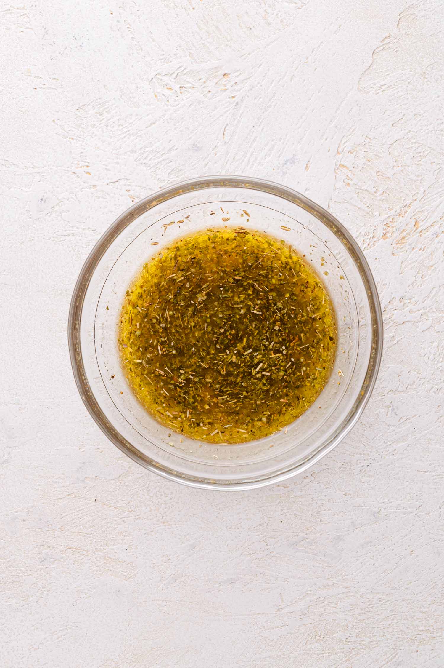 A clear glass bowl filled with a oil and seasoning mixture sits on a light textured surface.