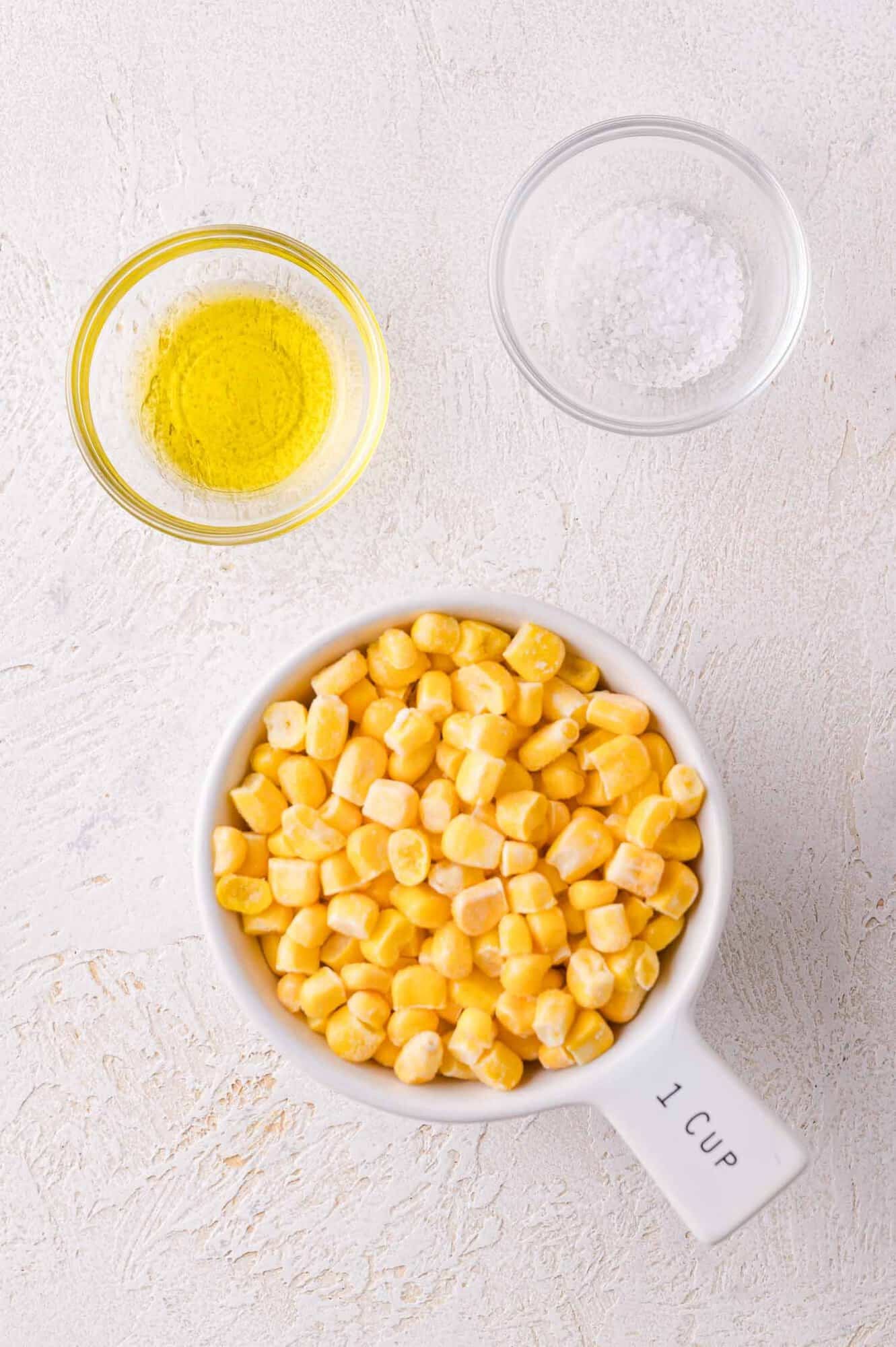 A measuring cup filled with corn kernels, a small bowl of olive oil, and a small bowl of salt on a light surface.