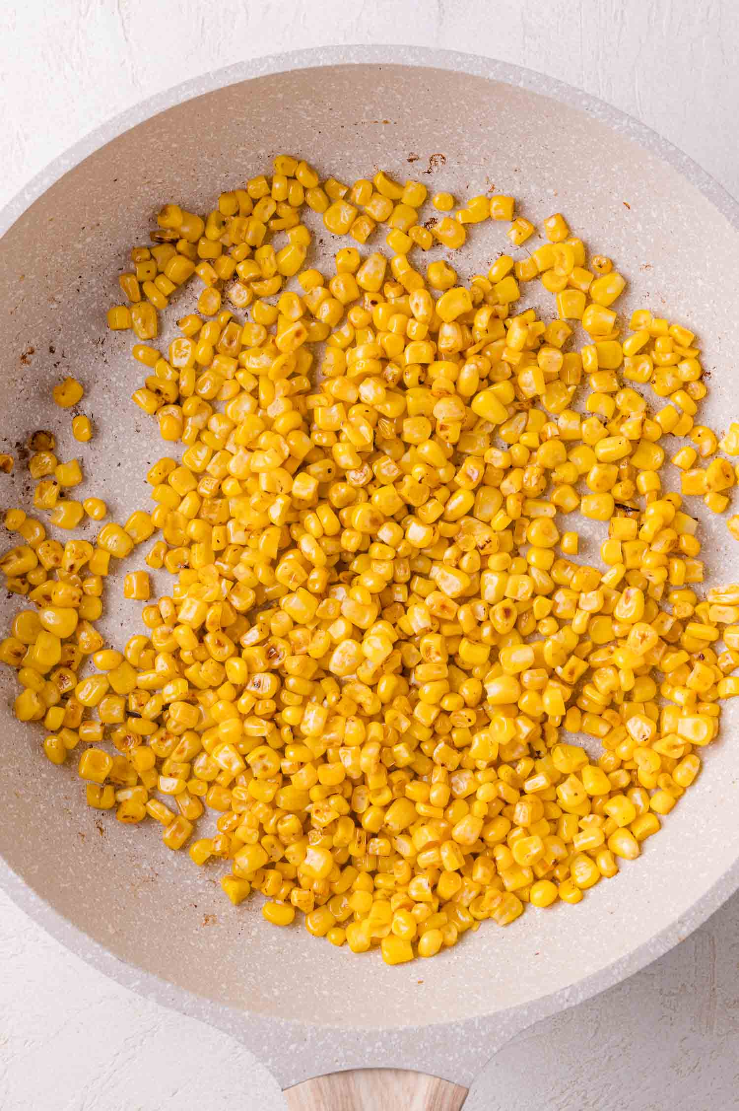 Corn kernels being sautéed in a light-colored frying pan on a white surface.