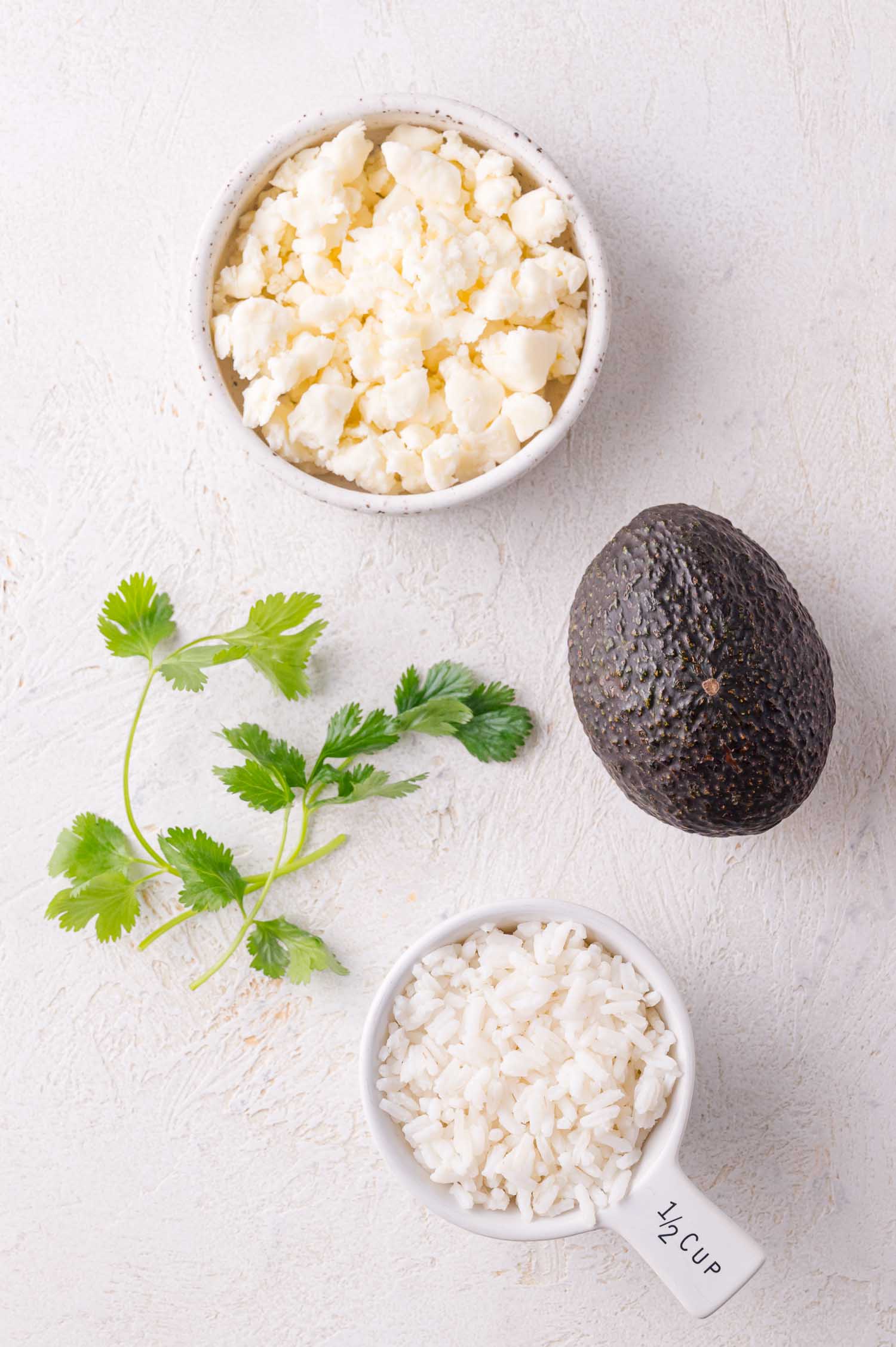 A bowl of crumbled cheese, a whole avocado, a sprig of cilantro, and a measuring cup with cooked rice on a white surface.