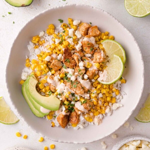 A bowl of rice topped with grilled corn, seasoned chicken pieces, avocado slices, lime wedges, crumbled cheese, and a light drizzle of sauce.
