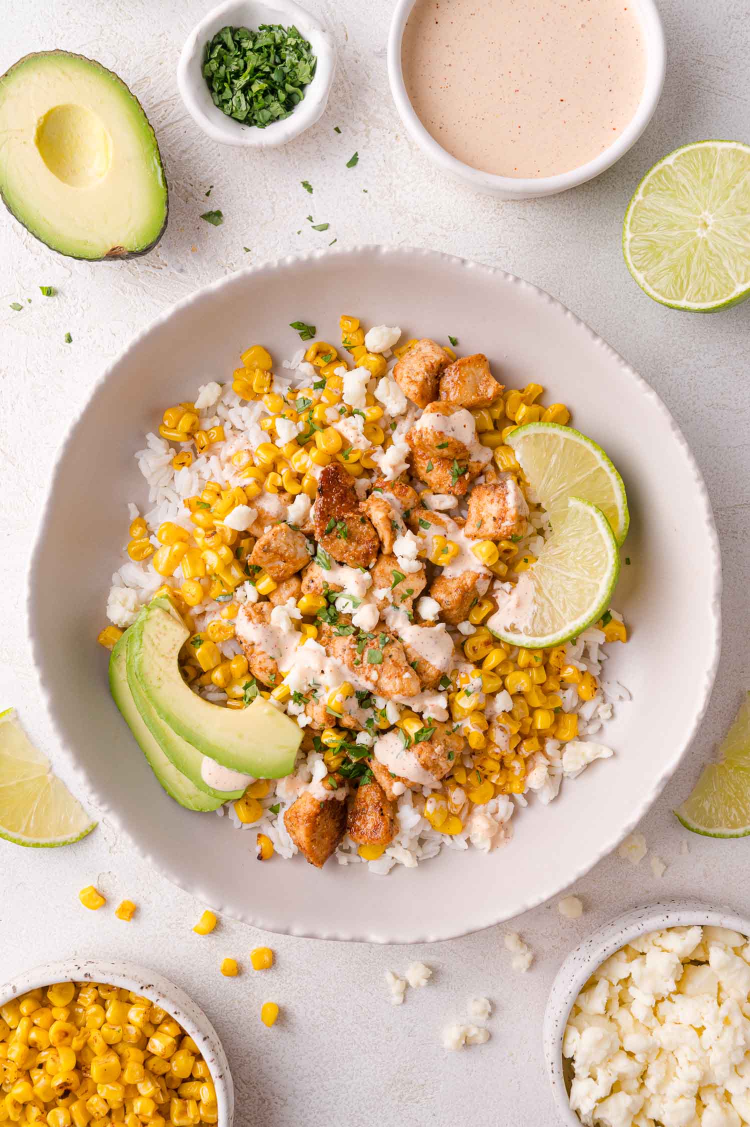 A bowl of rice topped with seasoned chicken, corn, avocado slices, lime wedges, and crumbled cheese, surrounded by small bowls of sauce, herbs, corn, and cheese.