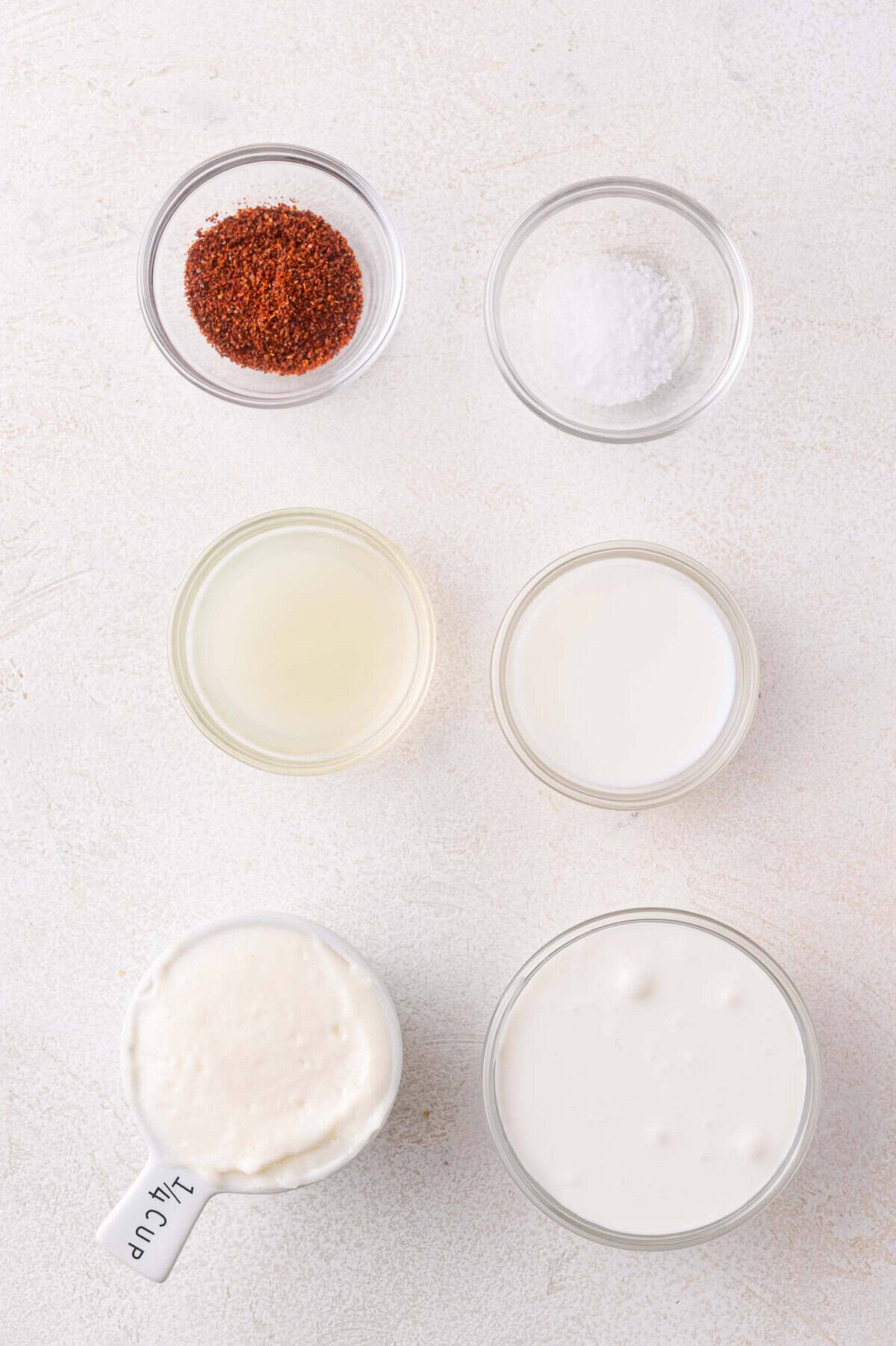 Six small bowls on a white surface containing chili powder, salt, lime juice, milk, mayonnaise in a 1/4 cup measure, and sour cream.
