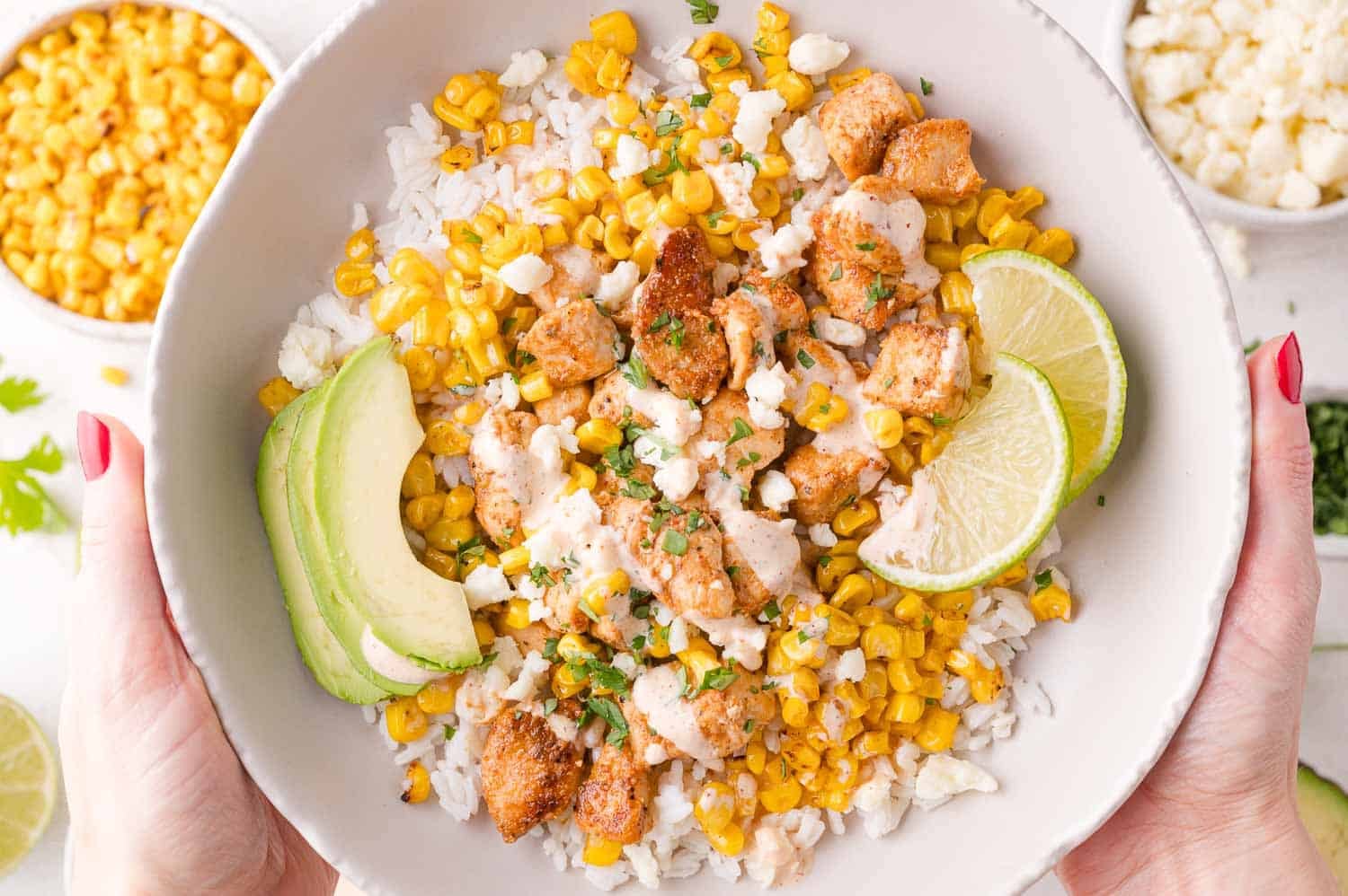 A bowl of rice topped with grilled chicken, corn, crumbled cheese, avocado slices, and lime wedges, held in two hands.