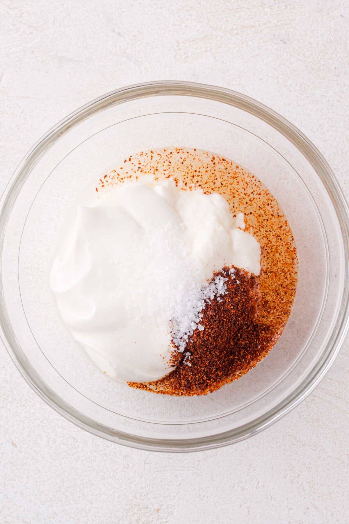 A glass bowl containing sour cream, chili powder, salt, on a light-colored surface.