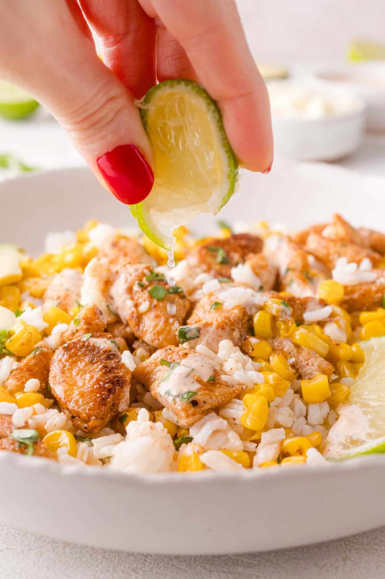 A hand squeezes a lime wedge over a bowl of rice, corn, and seasoned chicken pieces.