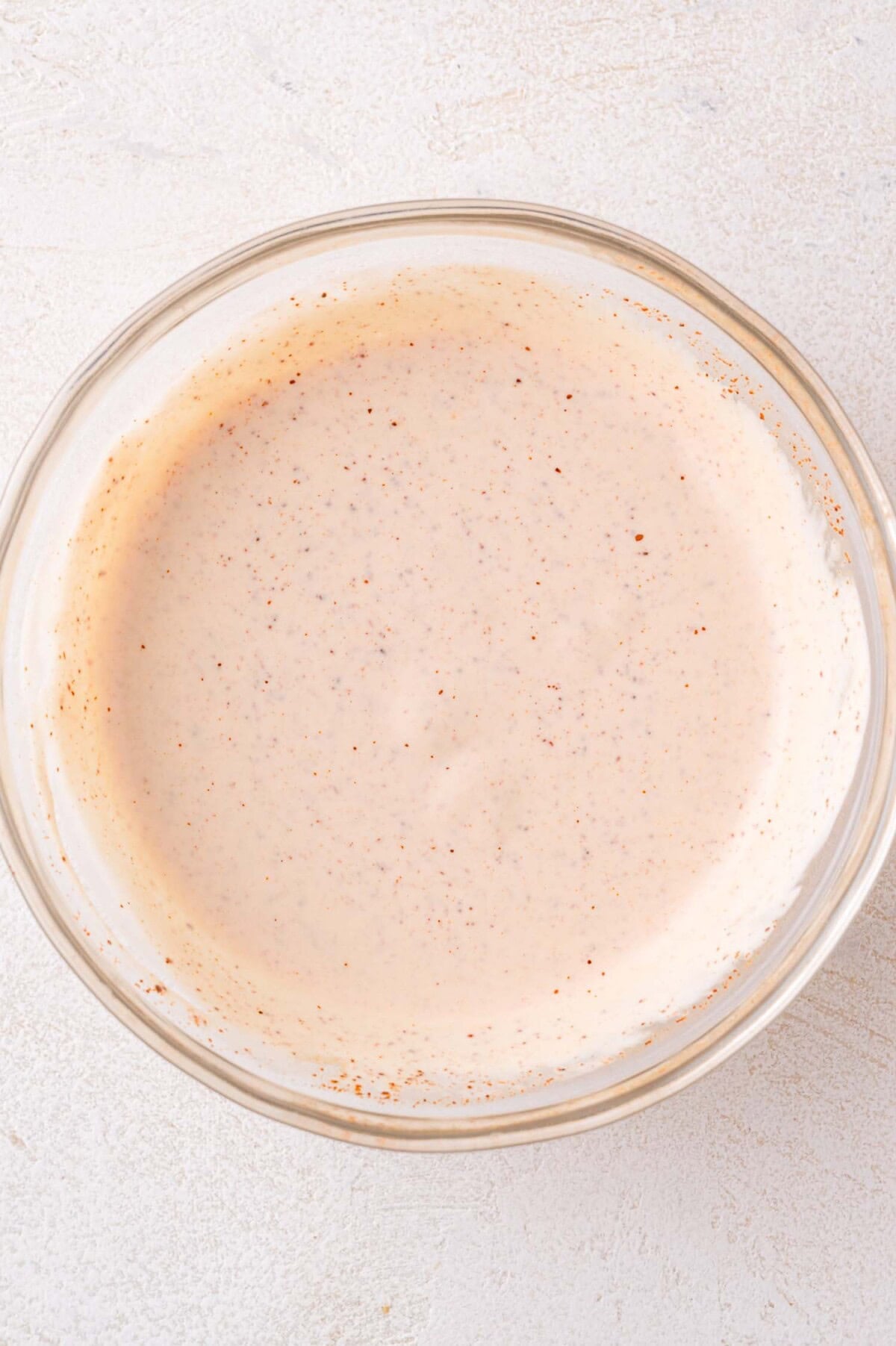 A clear glass bowl filled with a creamy, light orange sauce speckled with seasoning sits on a light-colored surface.