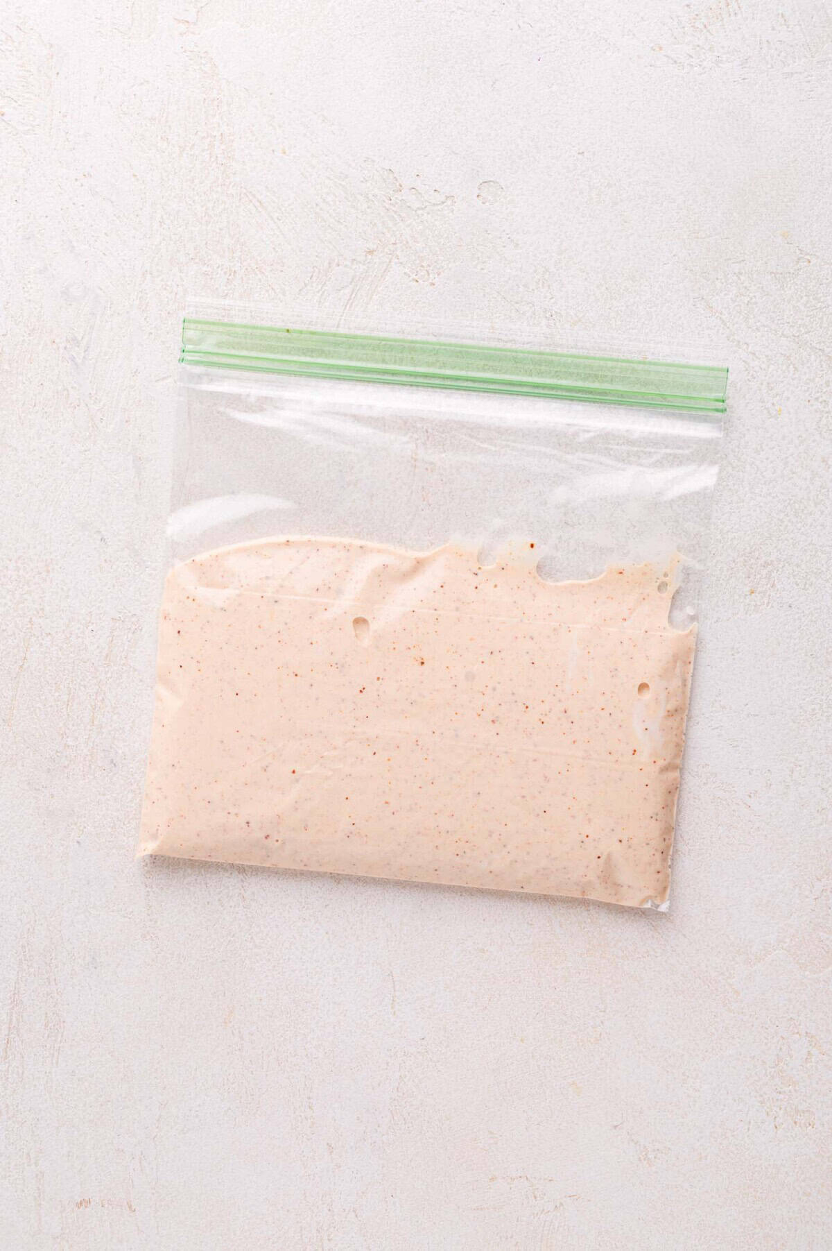 A sealed plastic zip-lock bag filled with a light beige, speckled sauce, placed on a light textured surface.