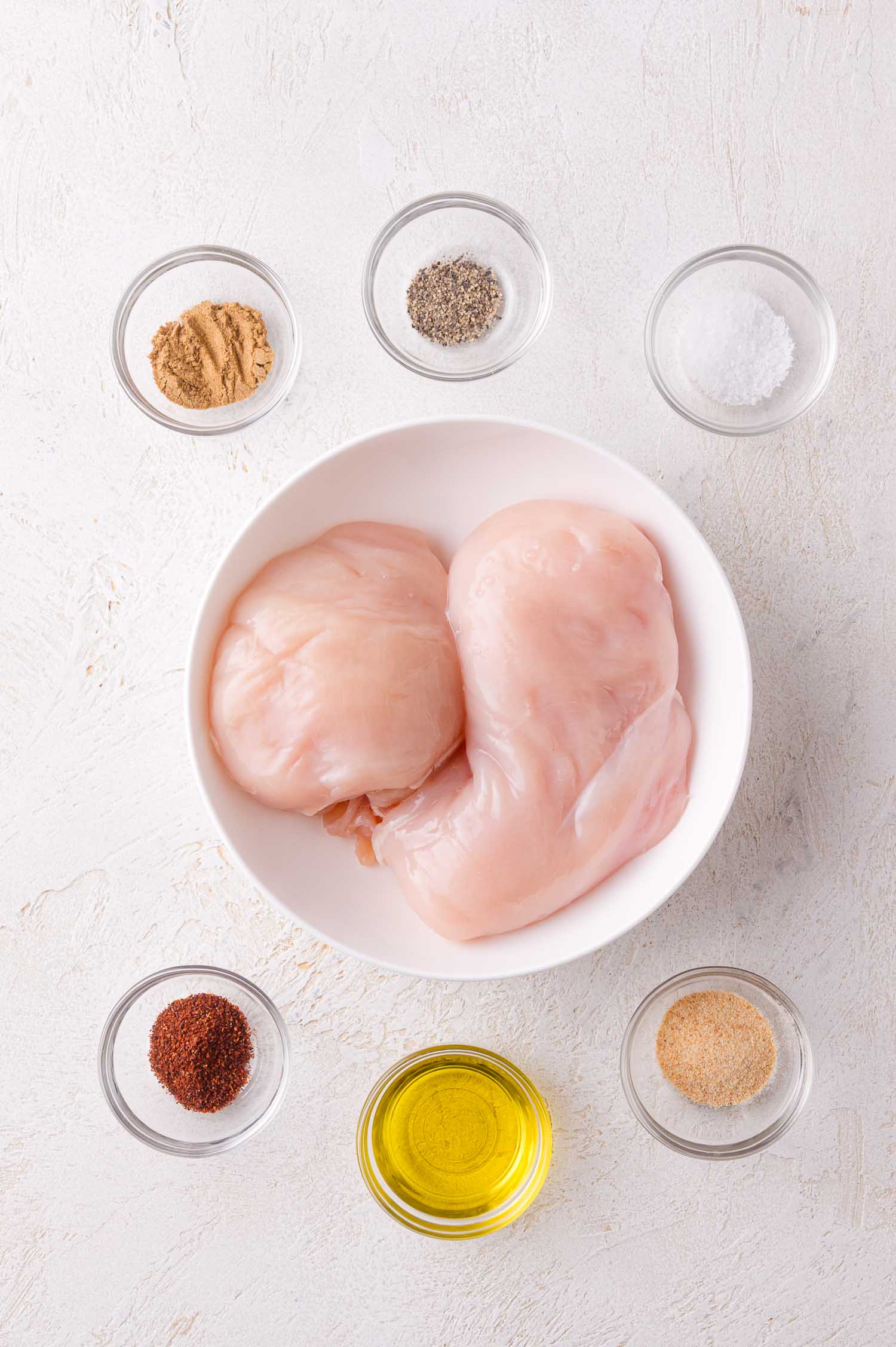 Raw chicken breasts in a white bowl, surrounded by small glass bowls containing oil, salt, pepper, ground spices, and seasonings on a white surface.