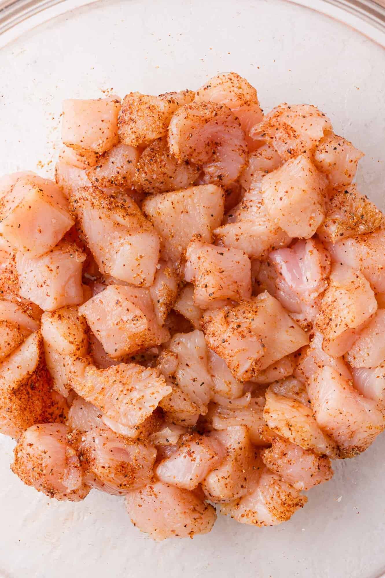 Raw diced chicken pieces coated with seasoning in a glass bowl.