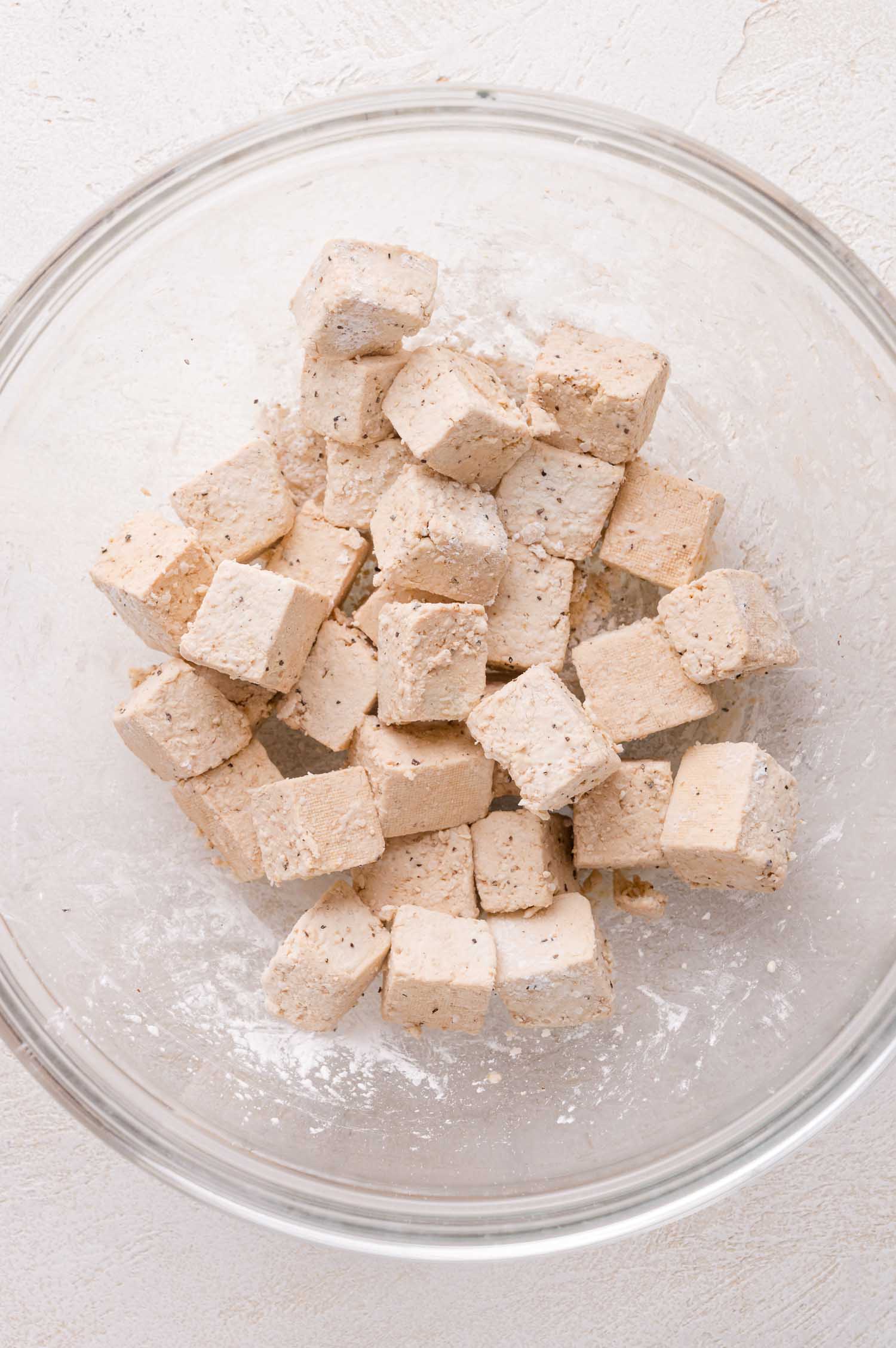 Cubed tofu coated in a light layer of seasoning and cornstarch sits in a clear glass mixing bowl on a white surface.