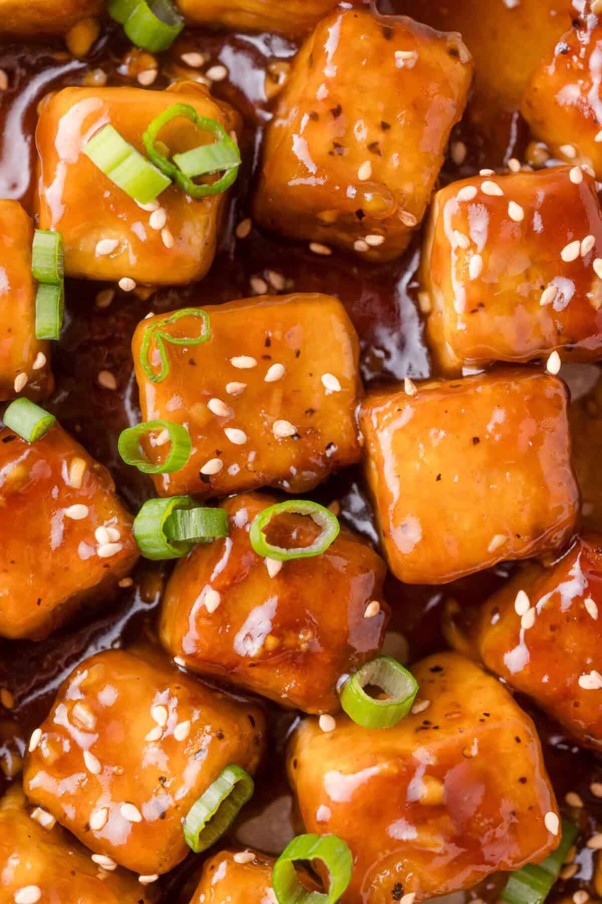 Close-up of glazed tofu cubes garnished with sliced green onions and sesame seeds, coated in a thick brown sauce.