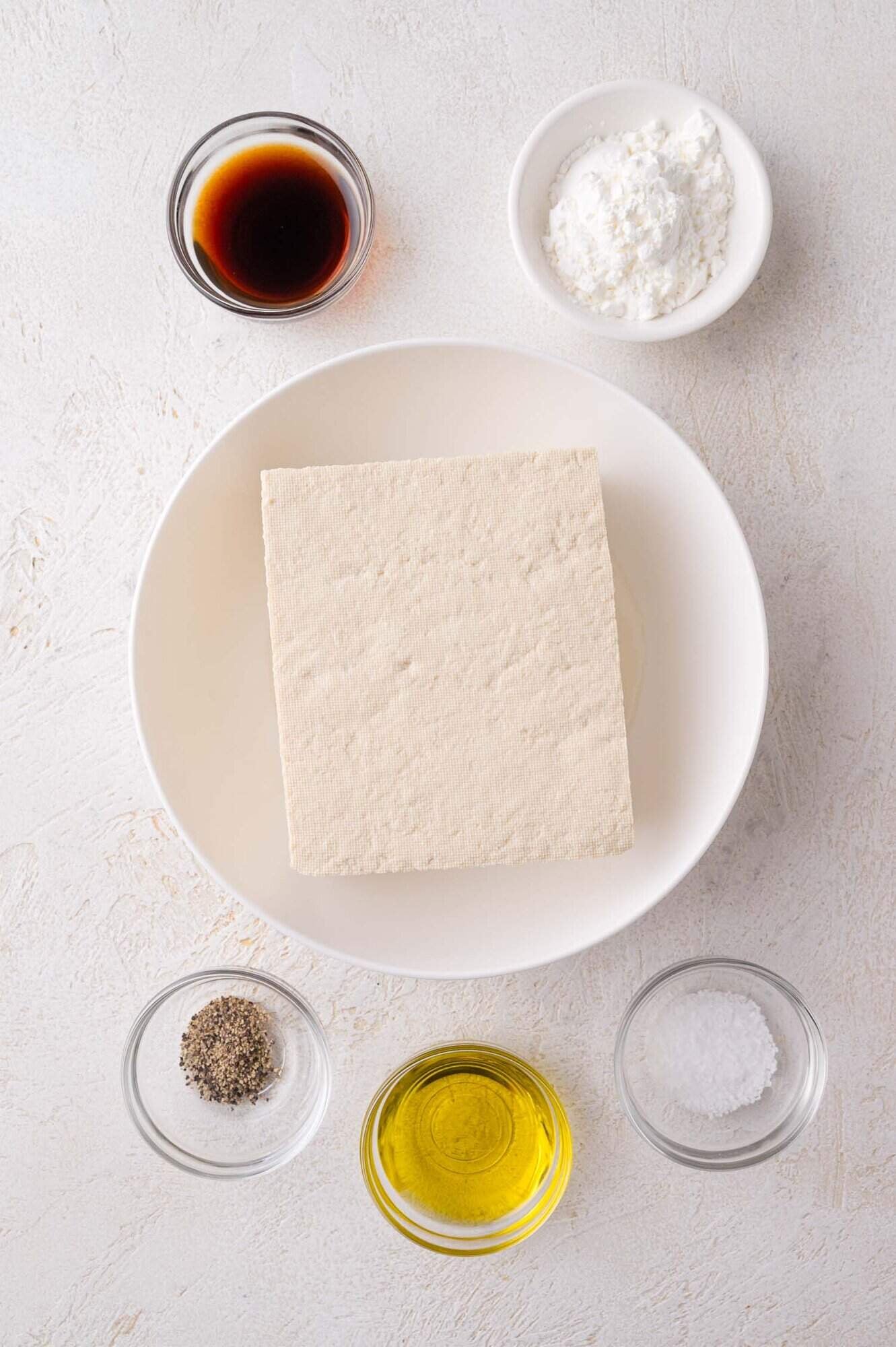 A block of tofu on a white plate surrounded by small bowls of soy sauce, cornstarch, ground pepper, olive oil, and salt.