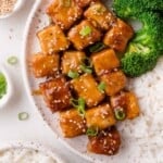 A plate with glazed teriyaki tofu cubes topped with sesame seeds and green onion, served with steamed broccoli and white rice.