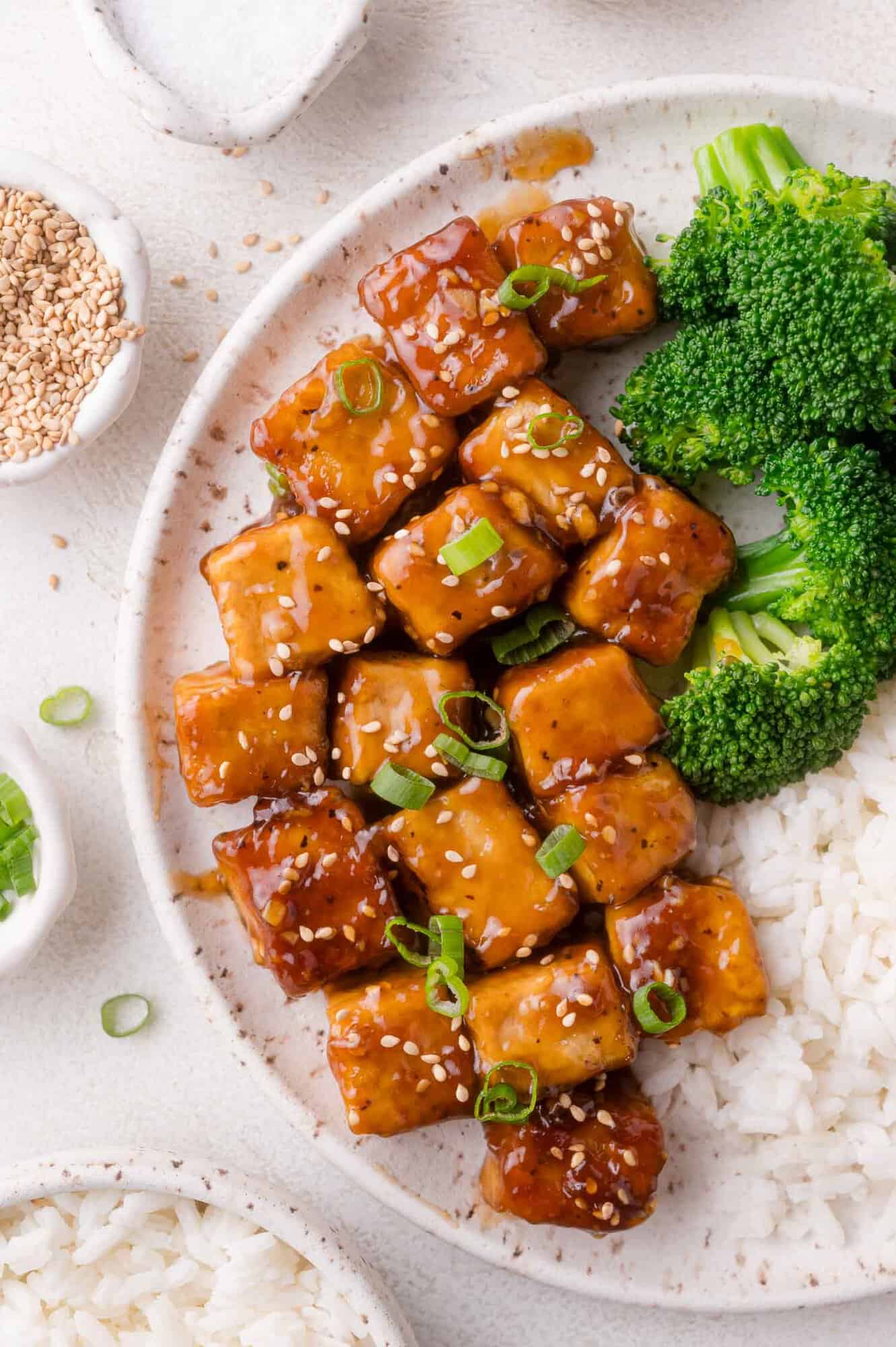 A plate with glazed teriyaki tofu cubes topped with sesame seeds and chopped green onions, served with steamed broccoli and white rice.