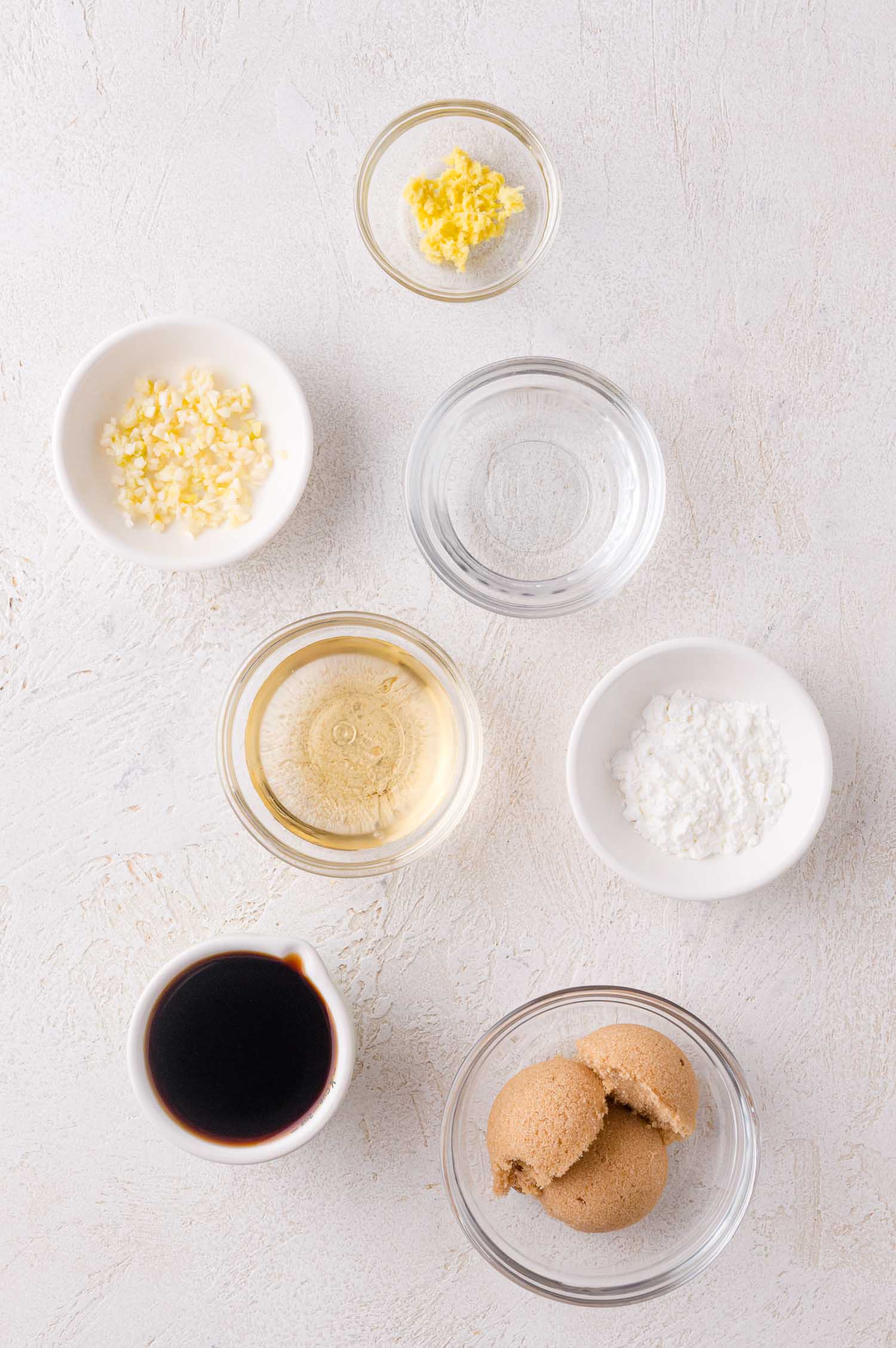 Seven small bowls containing minced garlic, minced ginger, water, rice vinegar, corn starch, soy sauce, and brown sugar arranged on a white surface.