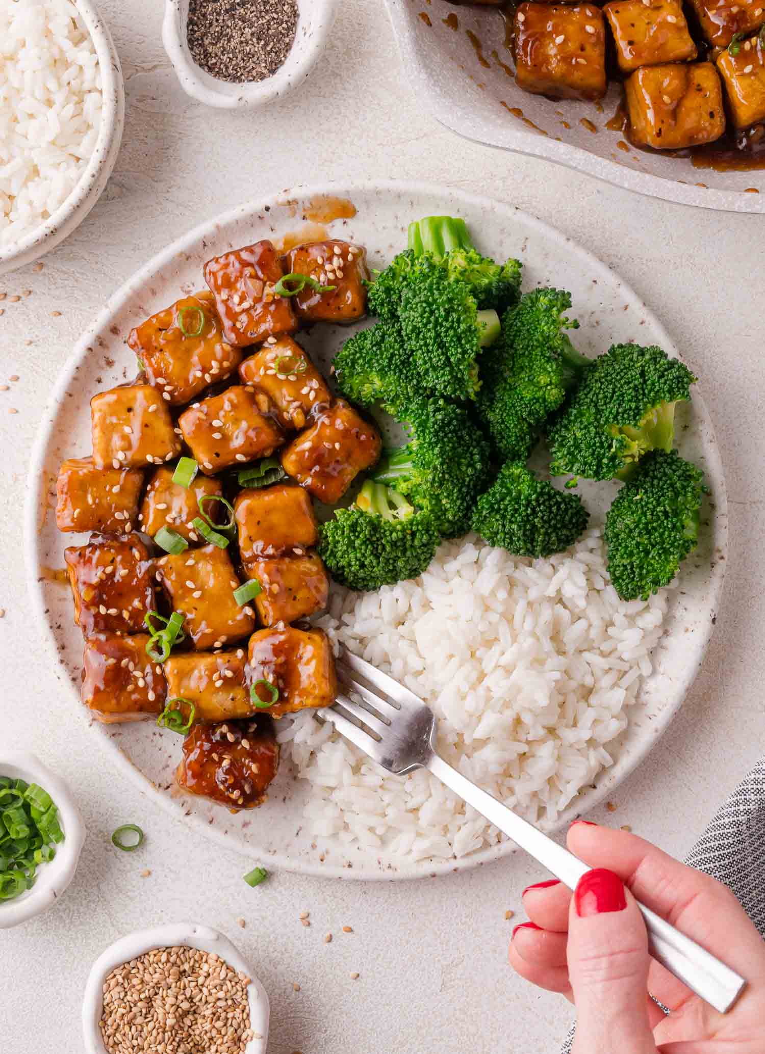 A plate with rice, broccoli, and teriyaki-glazed tofu cubes, with a hand holding a fork. Bowls of rice, pepper, green onions, and sesame seeds surround the plate.