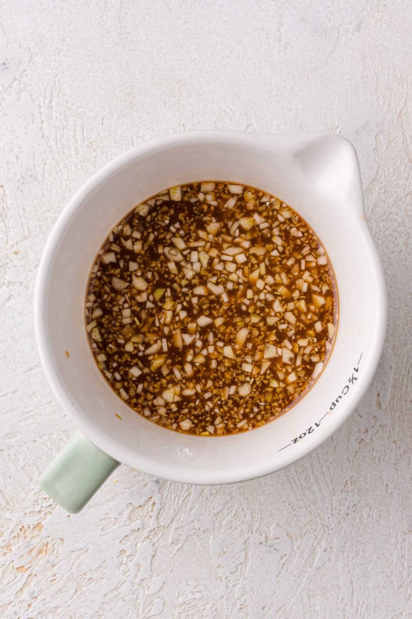 A white measuring cup contains a chopped garlic and soy sauce mixture on a light textured surface.
