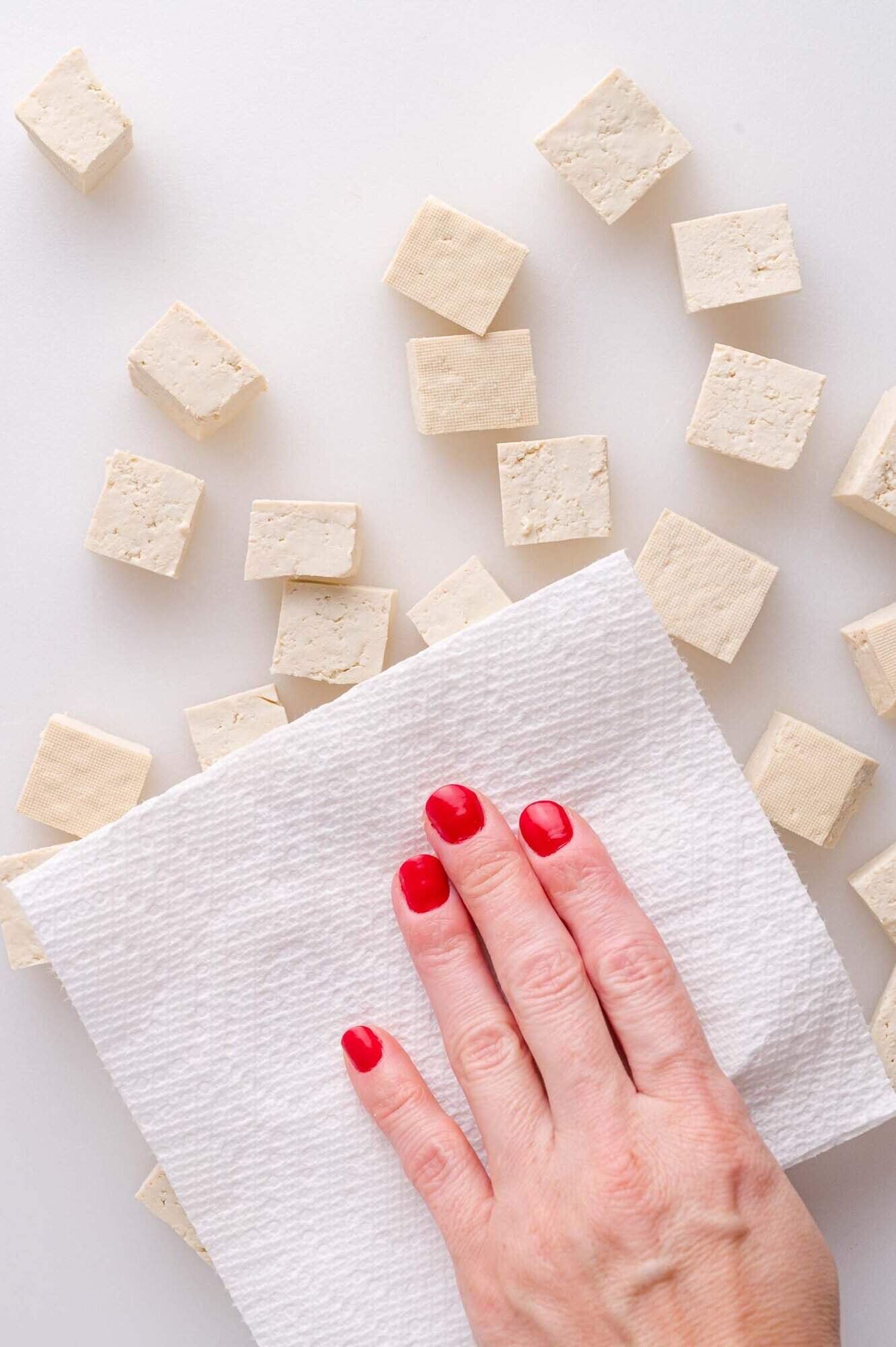 A hand with red nail polish presses a paper towel onto cubes of tofu on a white surface.