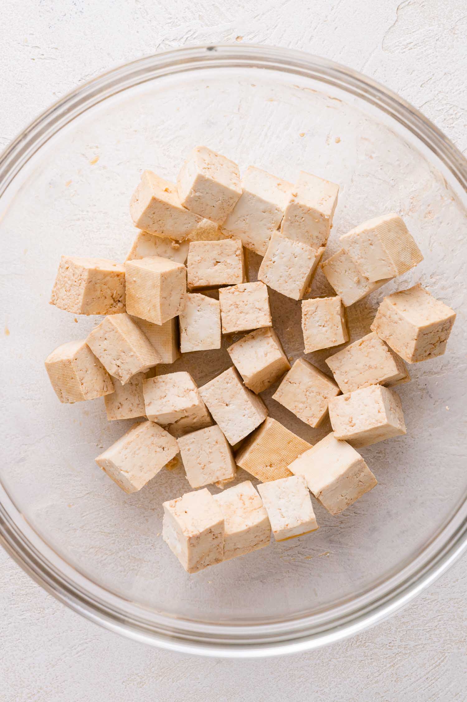 A clear glass bowl containing several cubes of plain tofu on a light-colored surface.
