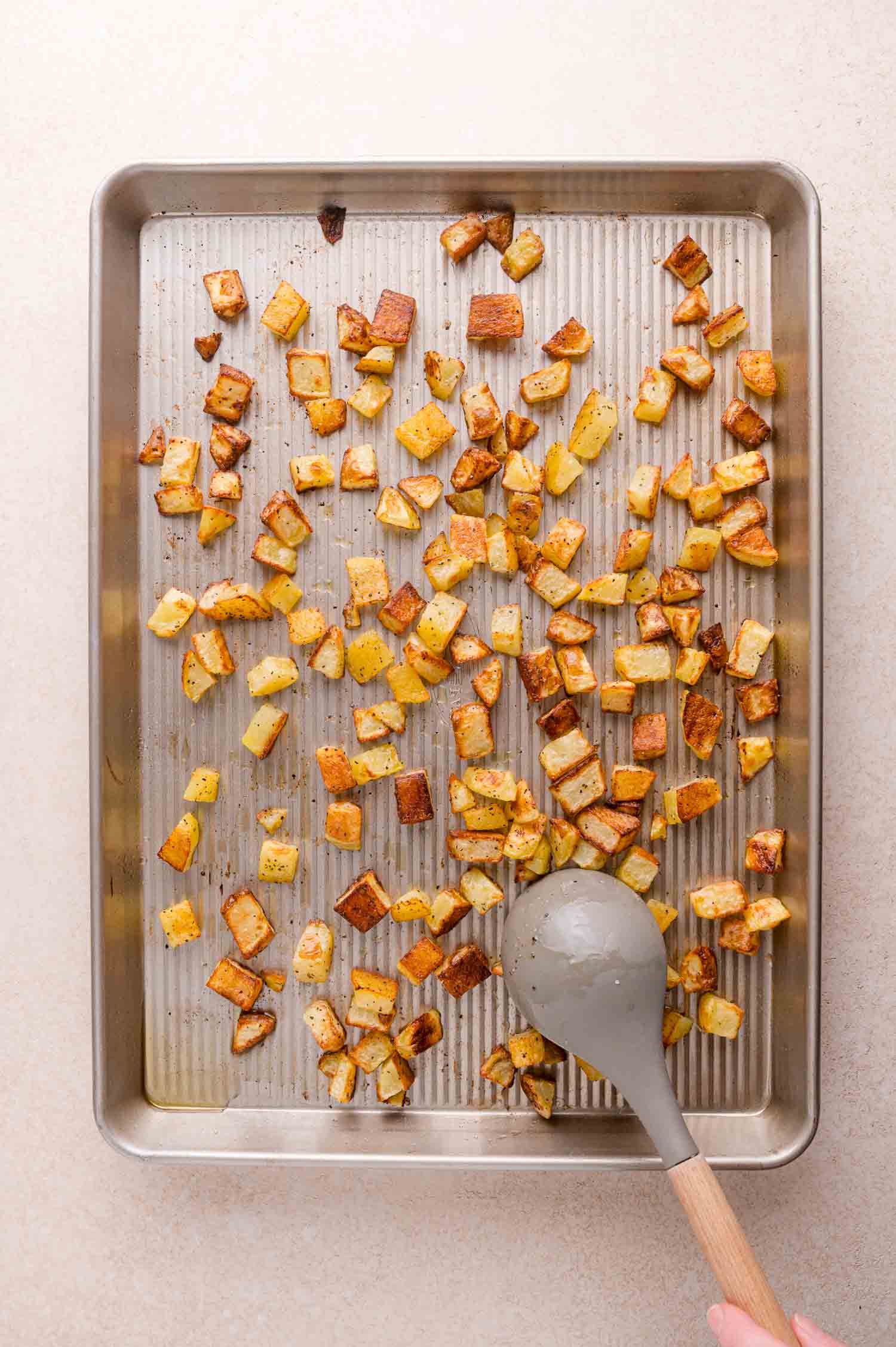 A baking tray with scattered roasted potato cubes, some browned, and a hand holding a gray spoon in the lower right corner.