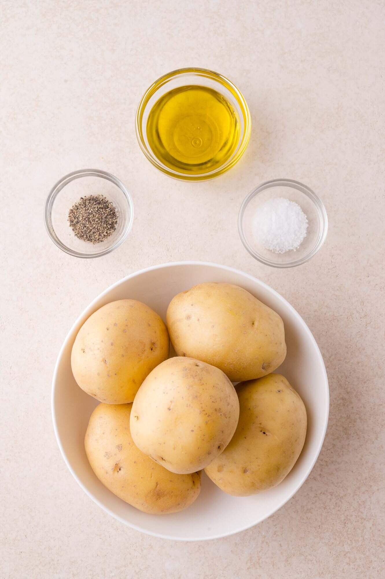 A bowl of whole potatoes next to small glass bowls containing olive oil, black pepper, and salt on a light surface.