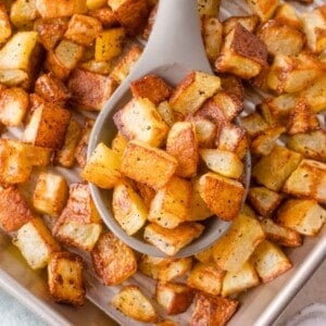 A baking tray filled with golden-brown roasted potato cubes, with a serving spoon lifting a portion from the tray.