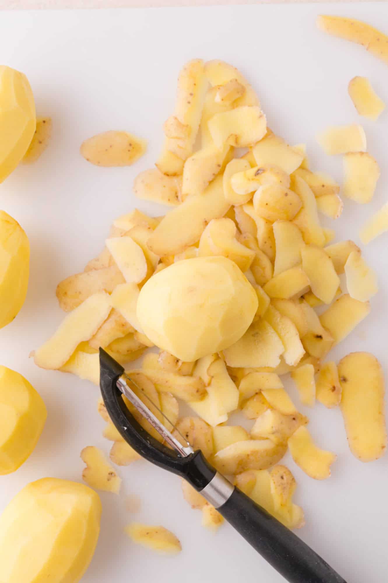 A pile of potato peels, several peeled potatoes, and a black vegetable peeler on a white surface.