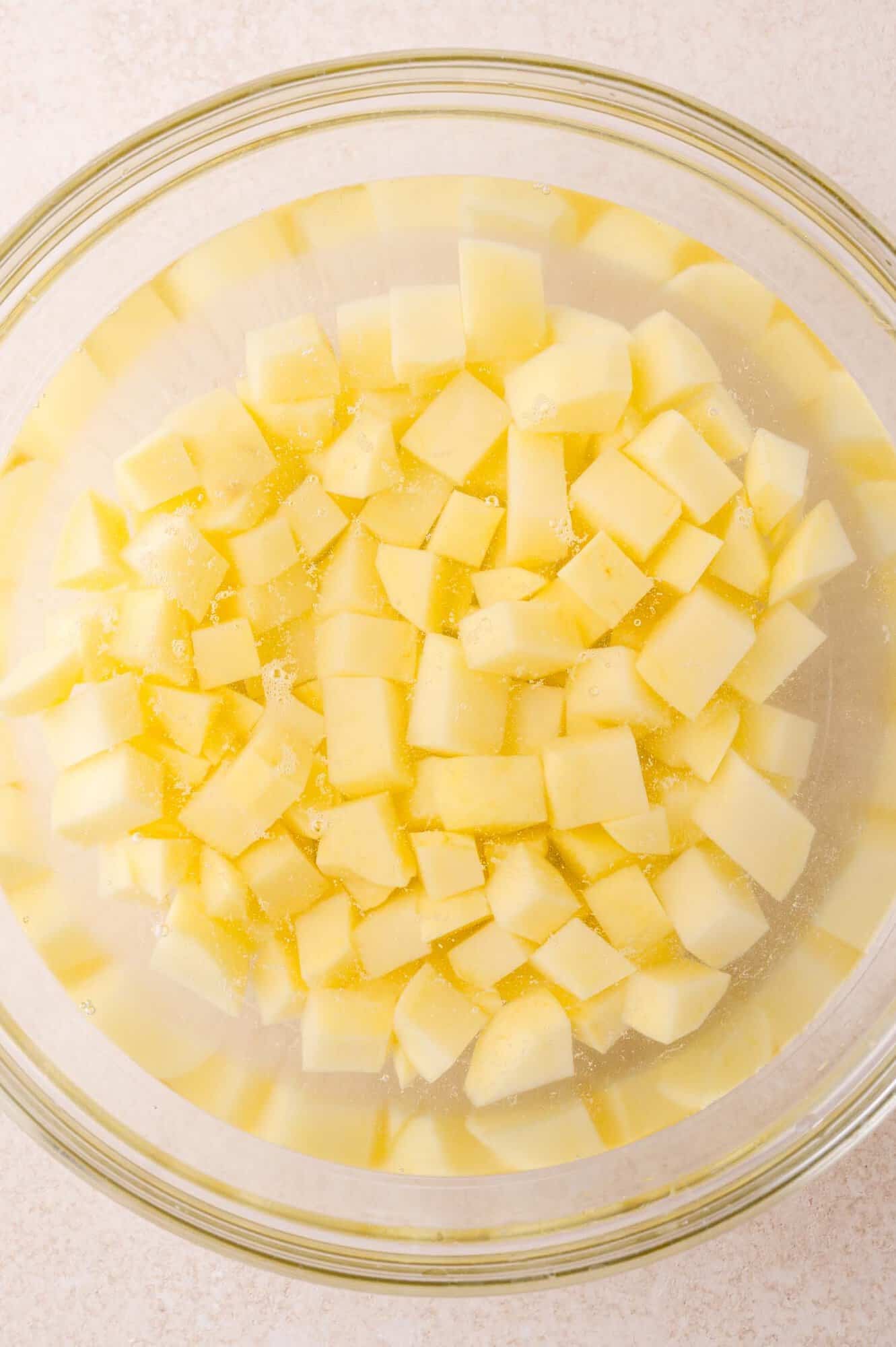 Cubed potatoes soaking in a glass bowl filled with water, viewed from above on a light-colored countertop.
