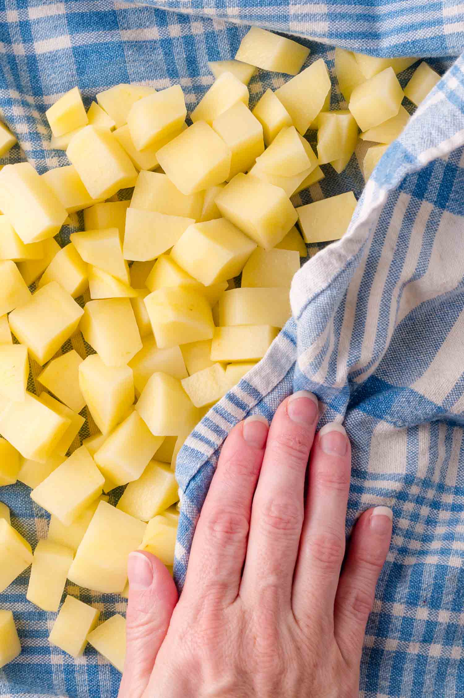 A hand drying diced potatoes with a blue and white checkered kitchen towel.