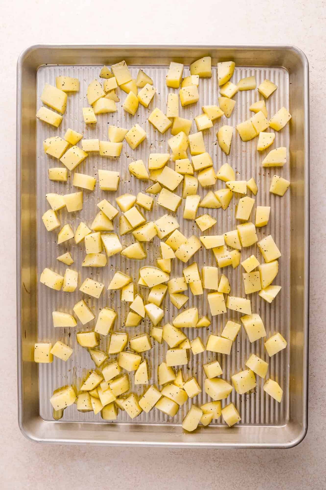 A baking sheet with evenly spread, diced potatoes seasoned with salt and pepper, ready for roasting.