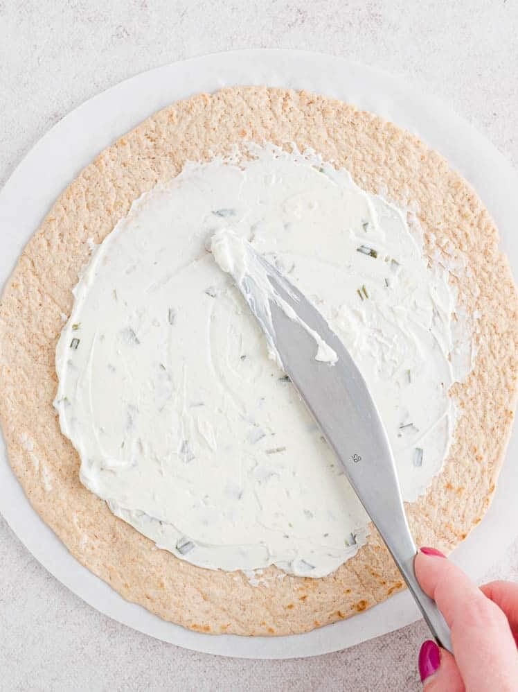 A hand using a knife to spread onion and chive cream cheese over a flour tortilla.