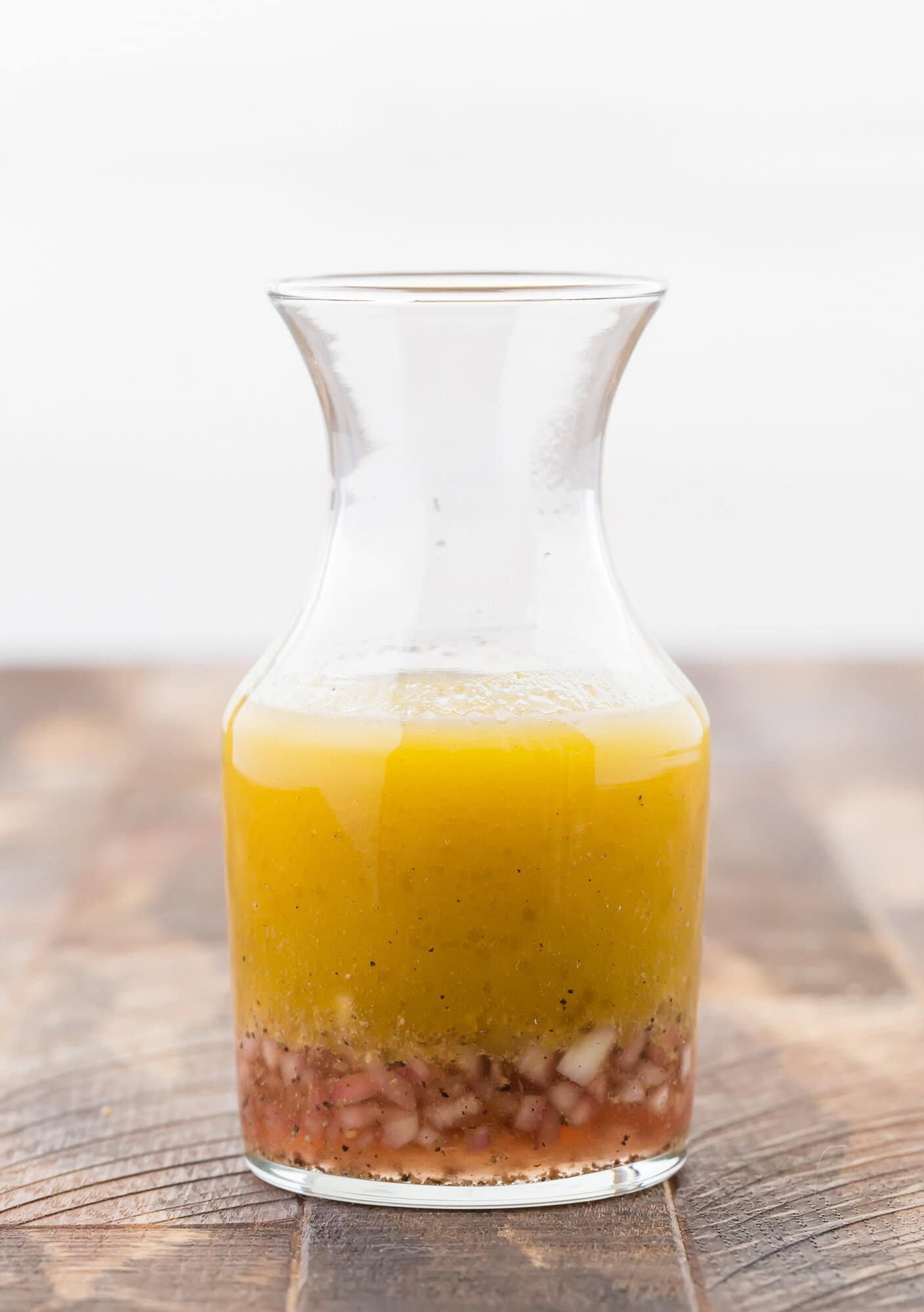 A clear glass carafe containing red wine vinaigrette with chopped shallots settled at the bottom, placed on a wooden surface.