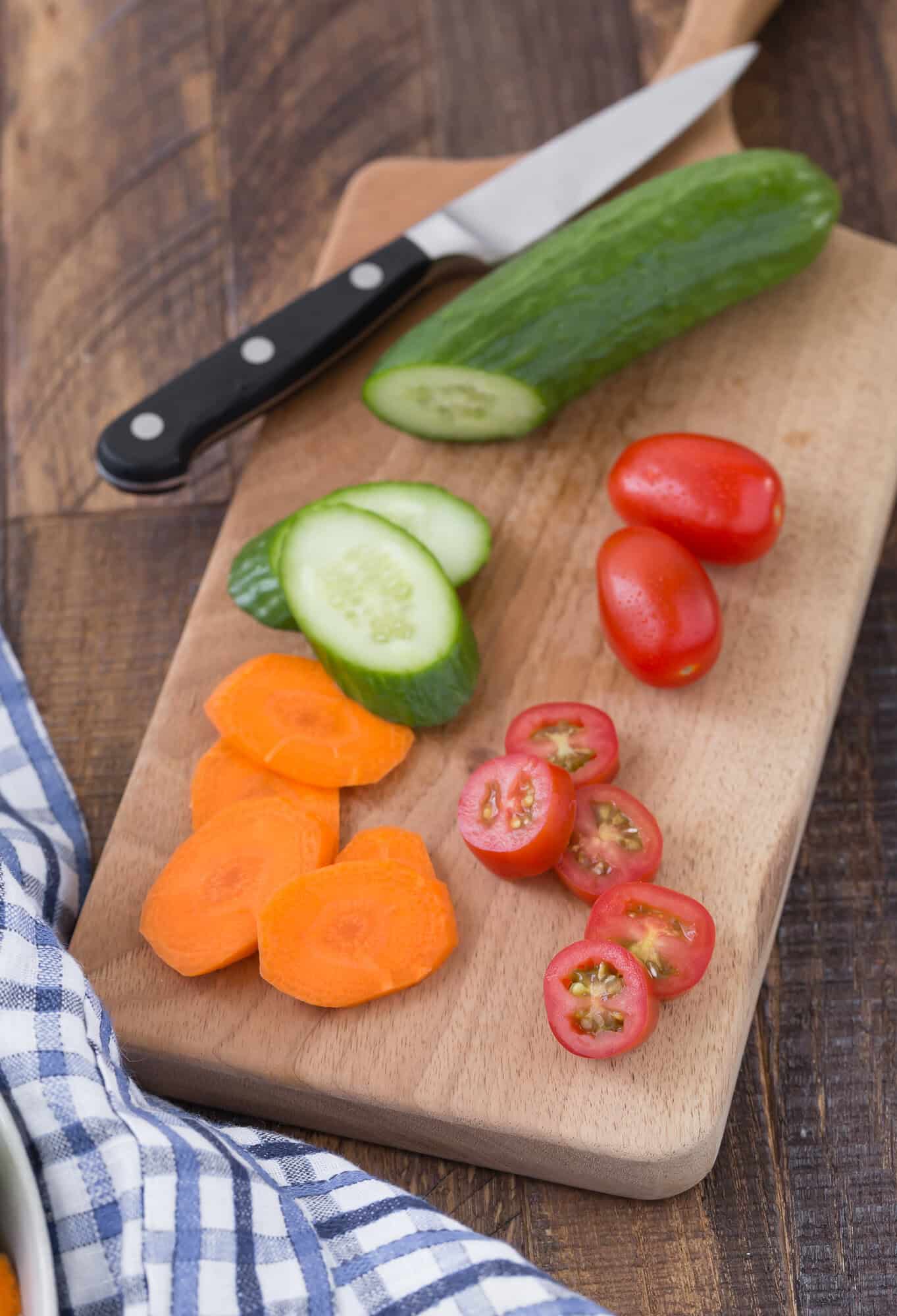 A cutting board with sliced cucumber, cherry tomatoes, and carrot, alongside a knife on a wooden surface and a blue-checkered cloth.