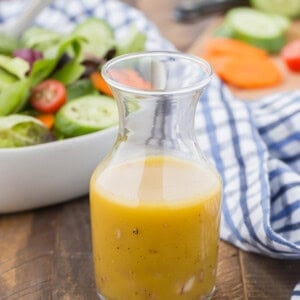 A glass carafe filled with red wine vinaigrette dressing sits on a wooden table next to a bowl of salad and a blue-and-white striped cloth.