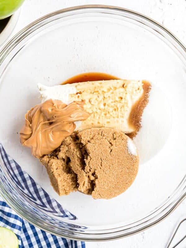 A glass bowl containing cream cheese, peanut butter, brown sugar, white sugar, and vanilla extract on a white surface.