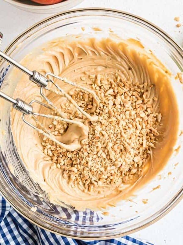 A glass bowl containing creamy batter with chopped nuts on top, partially mixed with an electric hand mixer.